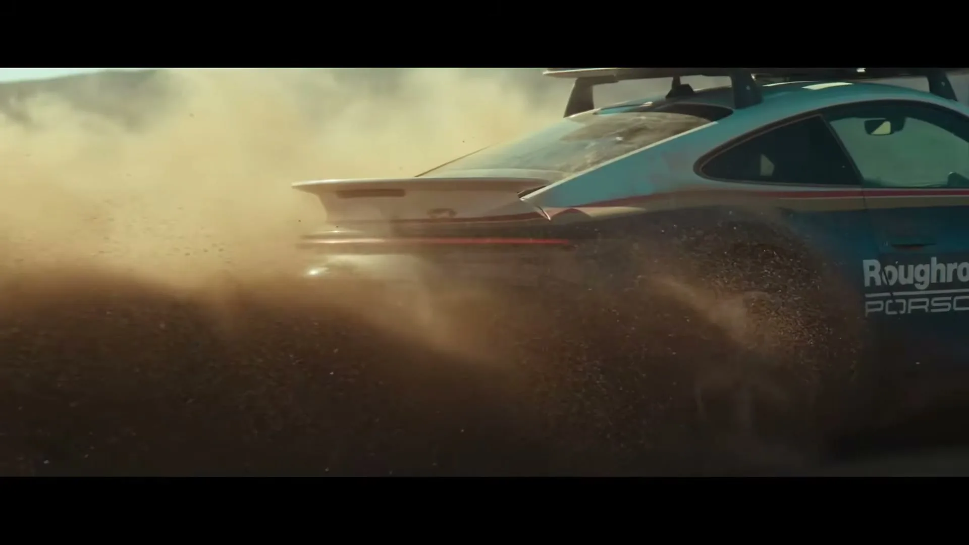A blue and white Porsche 911 Dakar kicks up a large cloud of dust as it speeds across a desert landscape during the day.