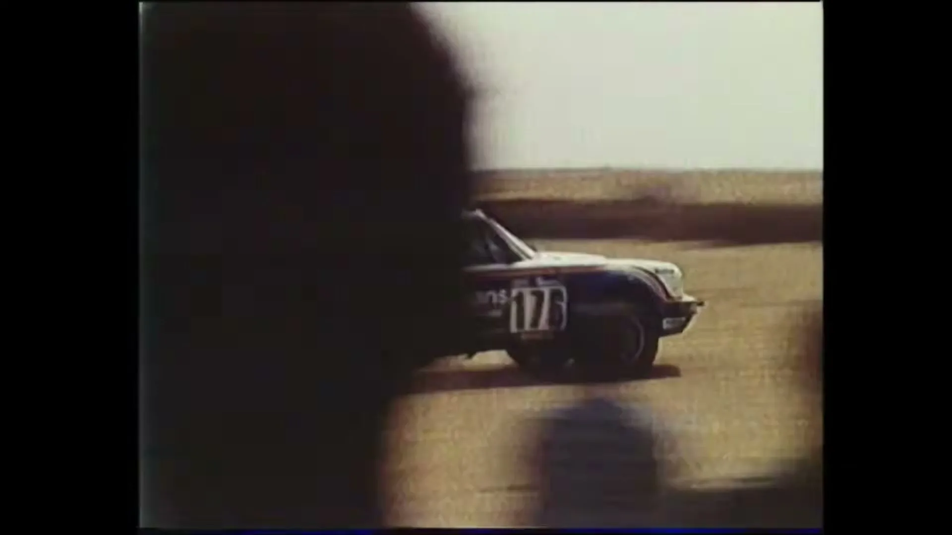 A vintage Porsche rally car with "Rothmans" livery speeds across a dusty track, as a crowd of people watch in the background.