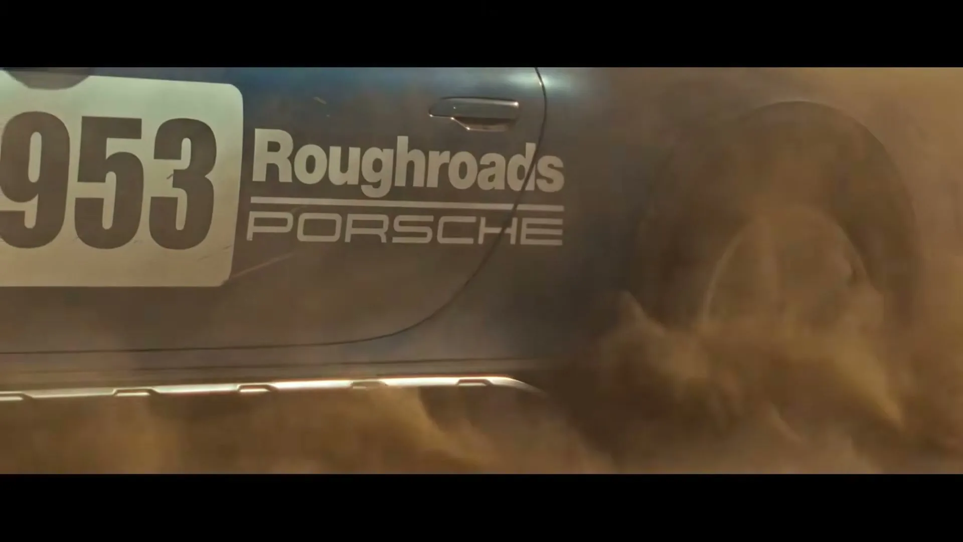 A blue and white Porsche 911 Dakar kicks up a cloud of dust as it drifts sideways on a dirt road in a desert landscape.