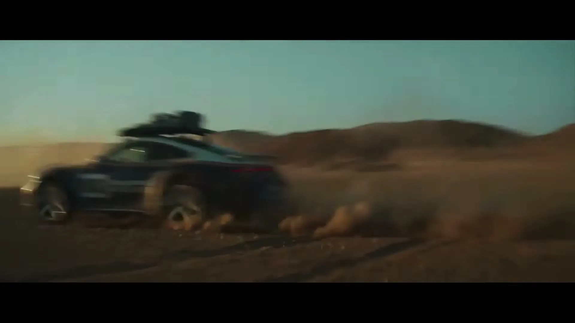 A black Porsche 911 Dakar drives fast through a dusty desert landscape, kicking up a large cloud of dust behind it under a clear sky.