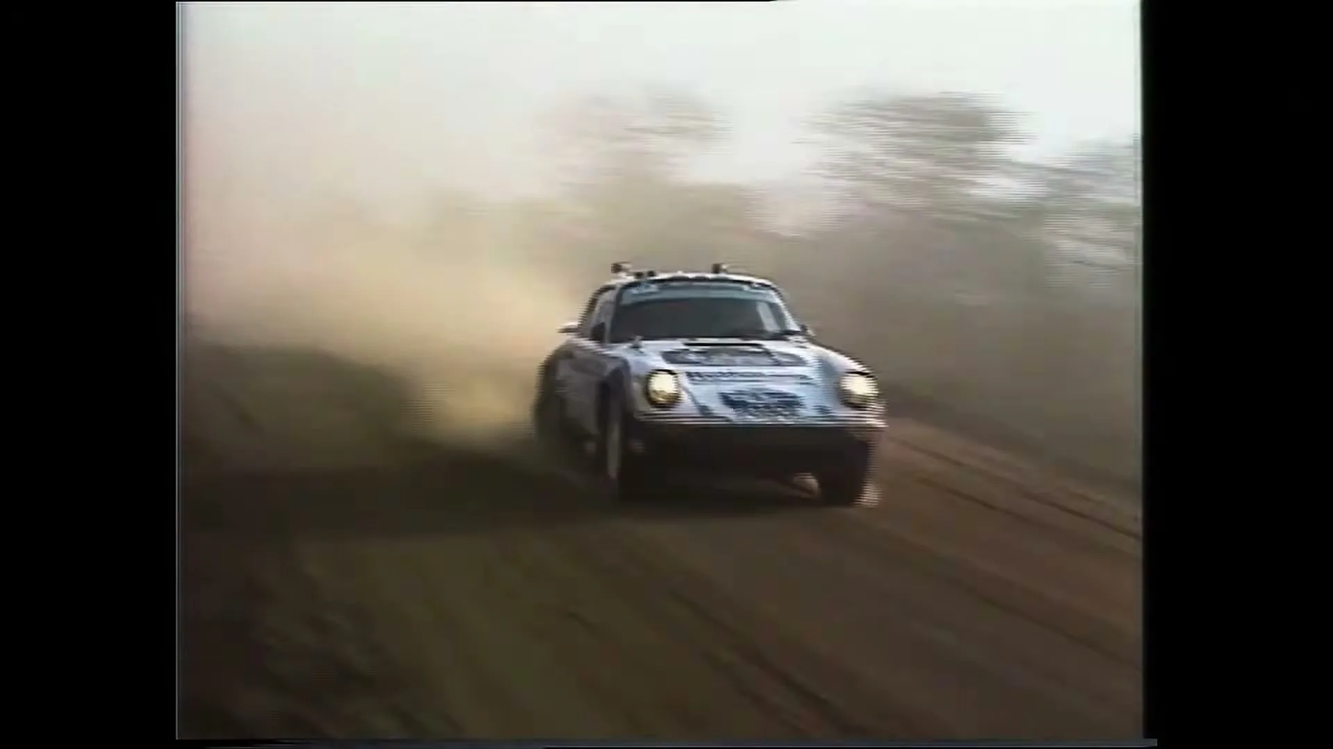 A white Porsche 911 Dakar rally car drives fast on a dusty dirt road, kicking up a large cloud of dust in a rural area.