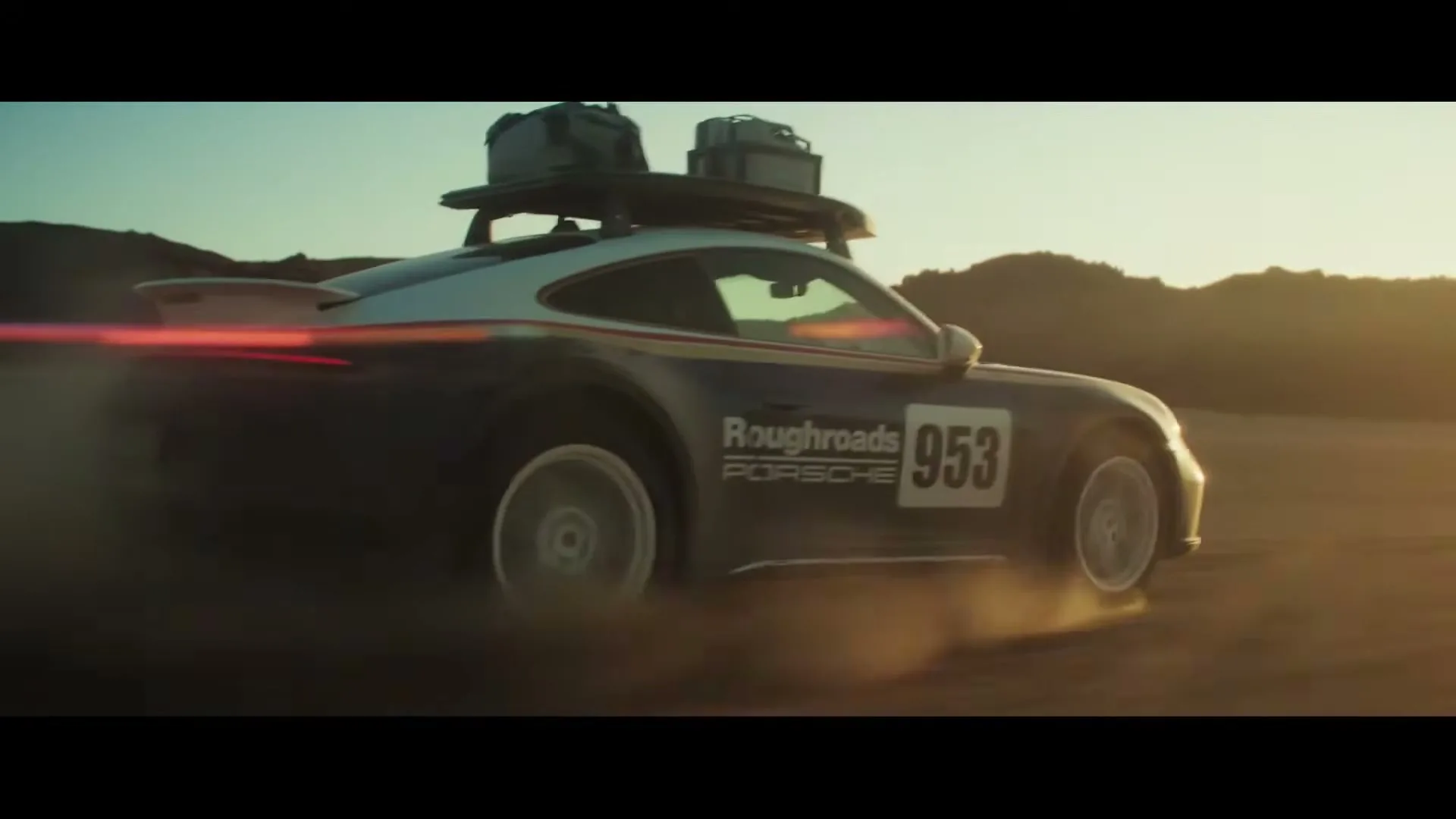 A dark blue Porsche 911 Dakar with a roof rack drives through a desert landscape, kicking up dust as the sun sets in the background.