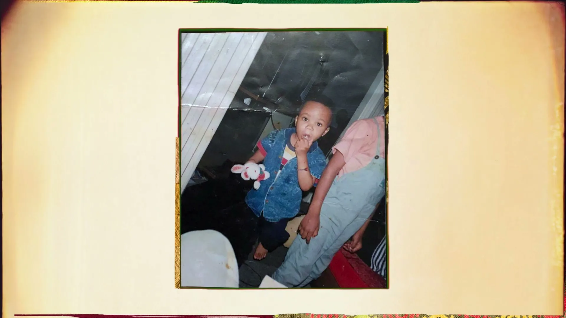 A young boy in a denim vest holds a toy bunny, standing next to an adult's leg in a faded photograph.