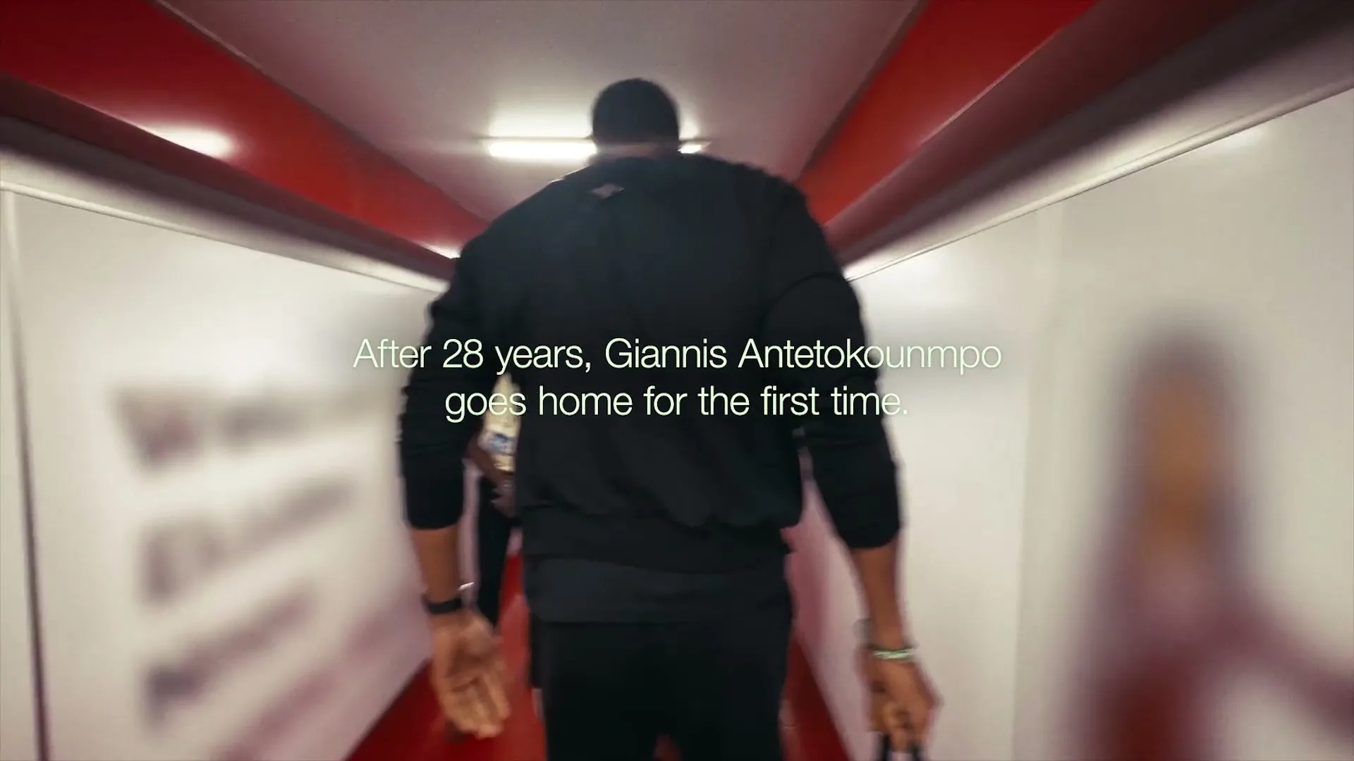 A tall man in a black tracksuit walks down a brightly lit airport jet bridge, carrying a duffel bag, with airport staff on either side.