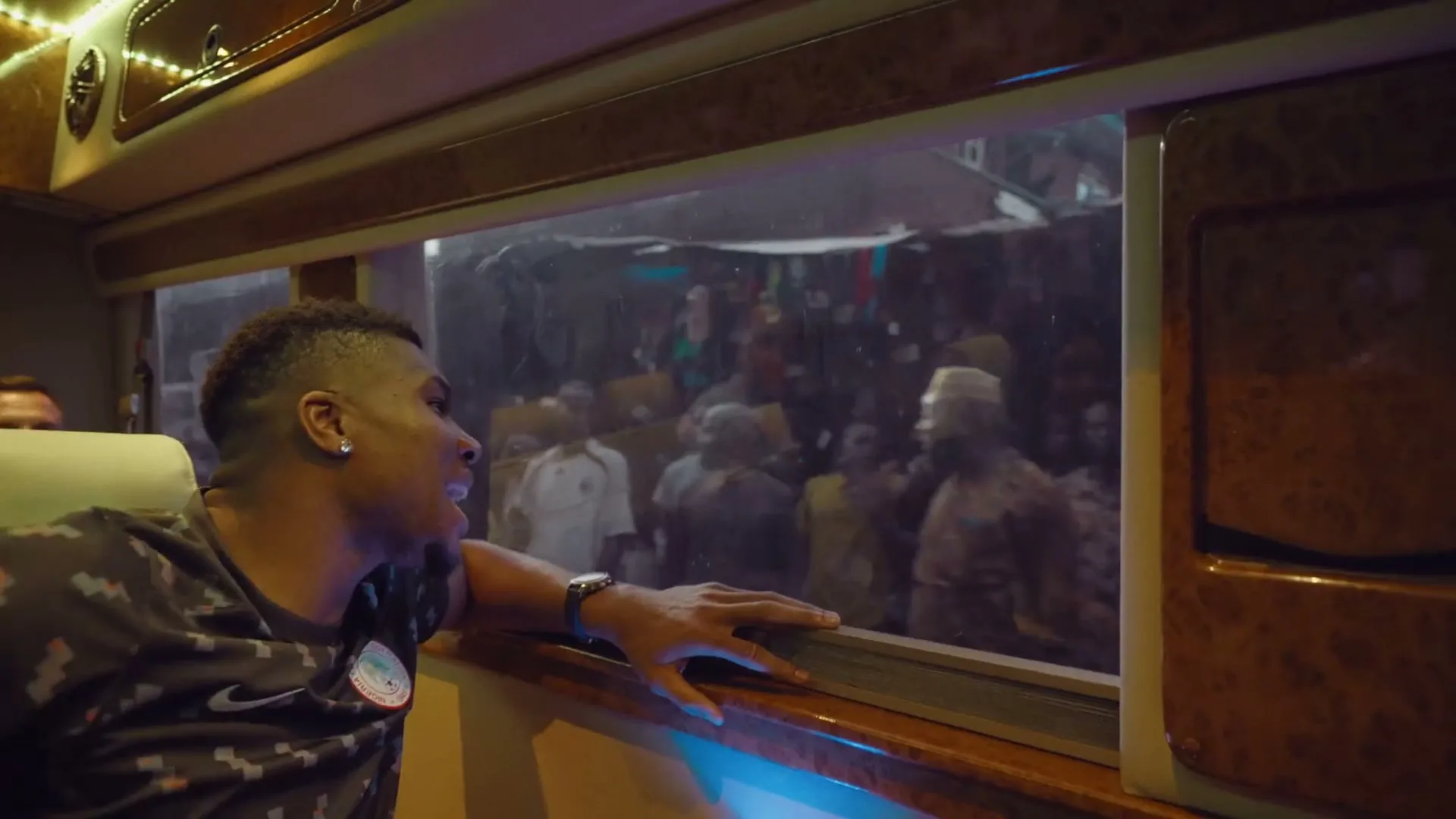 A young man in a patterned shirt looks out a bus window at a crowd of people on a busy street at night.