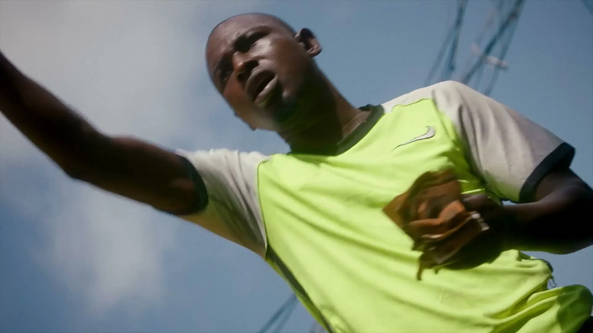 A man in a lime green and white shirt looks up at the sky, holding a brown object in his hand, with power lines in the background.
