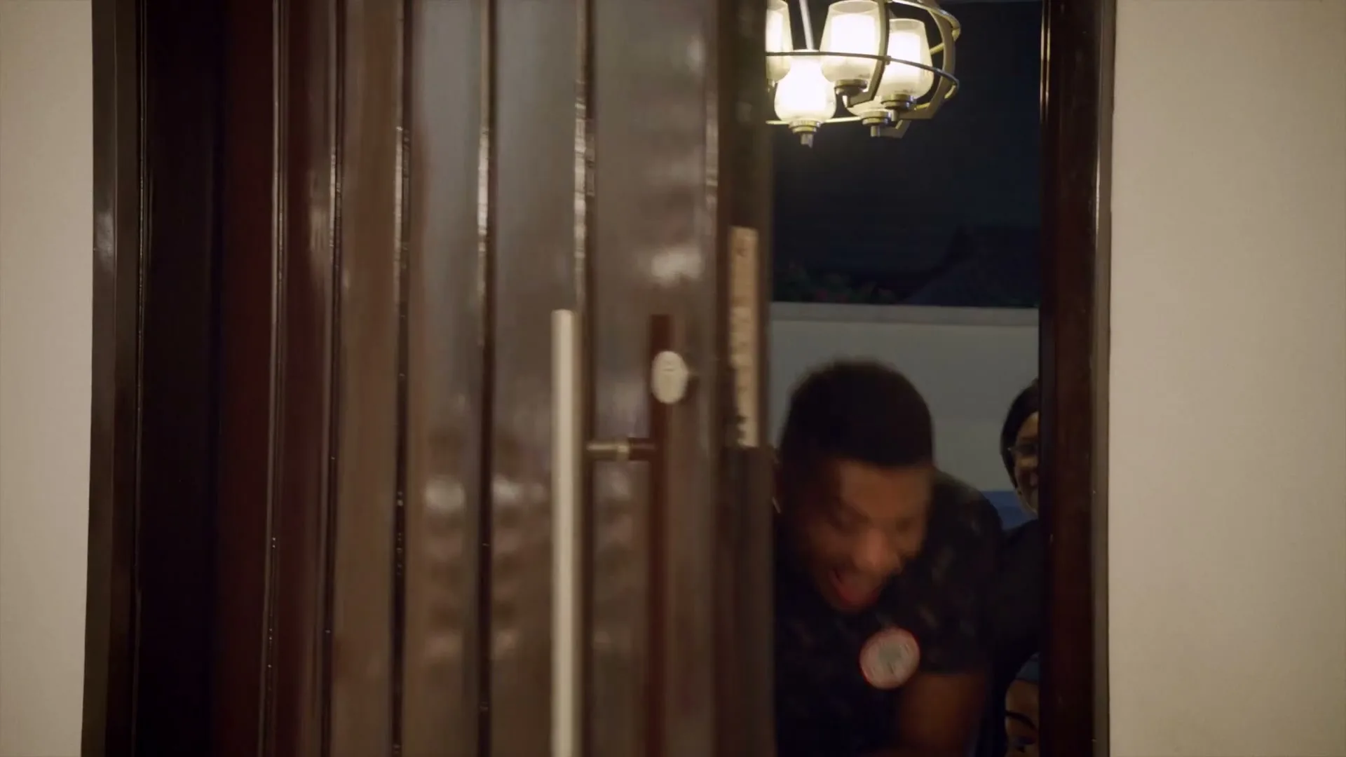 A young man in a dark patterned shirt peeks from behind a partially open wooden door, then steps into the doorway, smiling broadly.