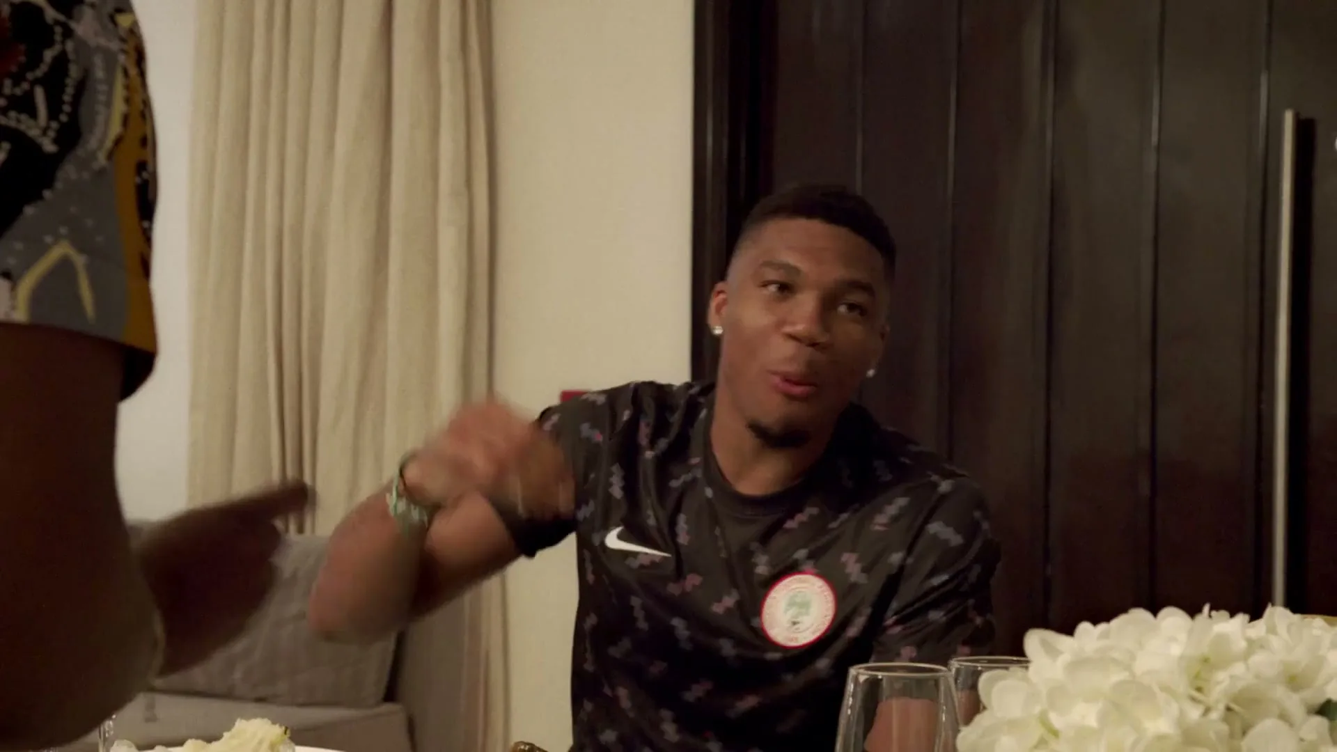 A young man in a black patterned t-shirt sits at a dining table, gesturing with his hands while talking to someone off-screen in an interior setting.