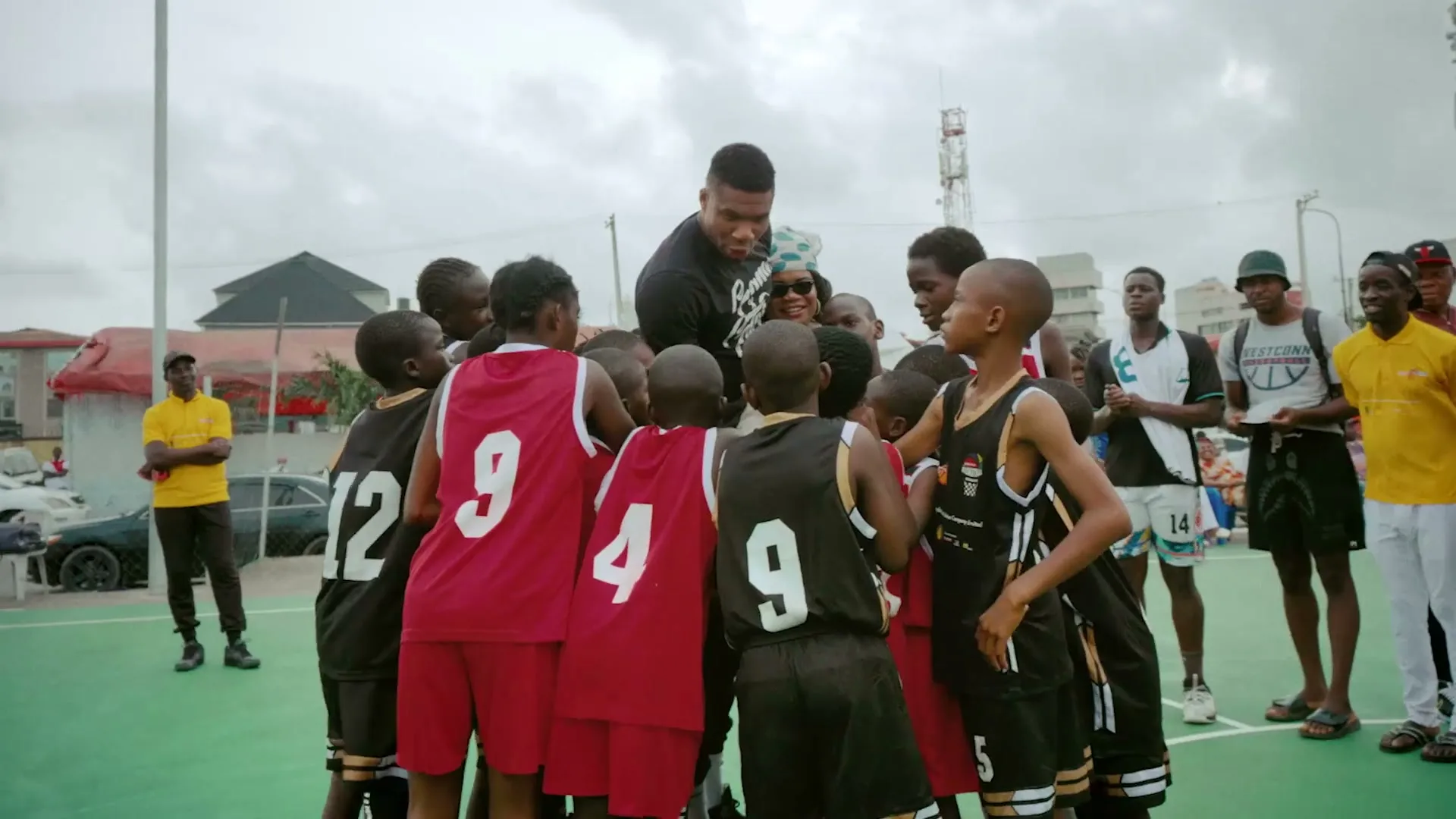 A group of young basketball players in red and black jerseys huddle around a male coach on an outdoor court, with other people watching in the background.