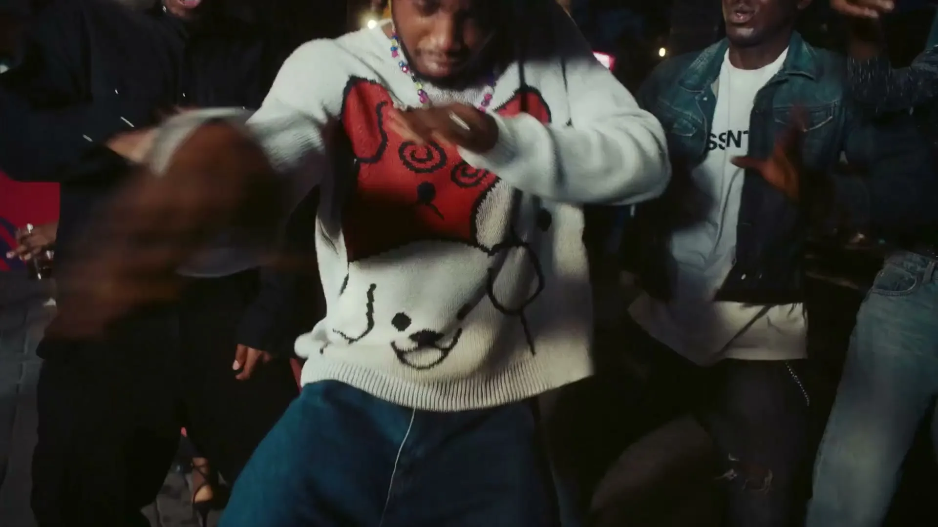 A young Black man in a white sweater with dog faces dances energetically in a crowded, dimly lit club.