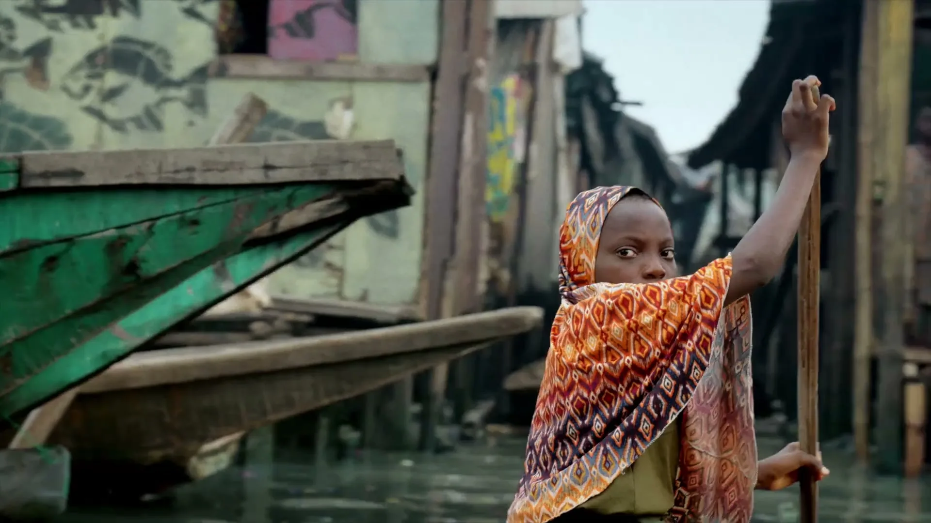 A young girl in a colorful headscarf paddles a boat, looking directly at the camera in a bustling waterside community.