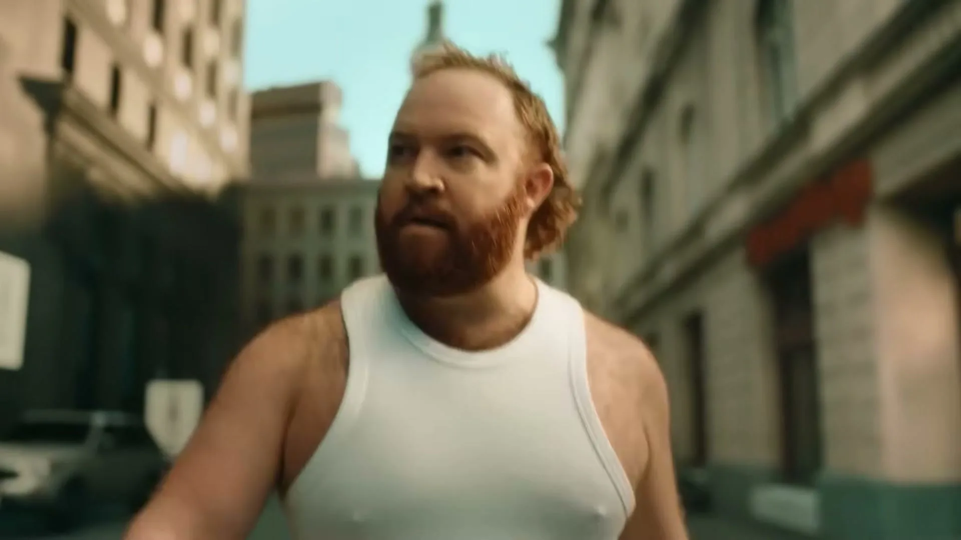 A man with a red beard and white tank top walks down a city street, looking around with a curious expression.