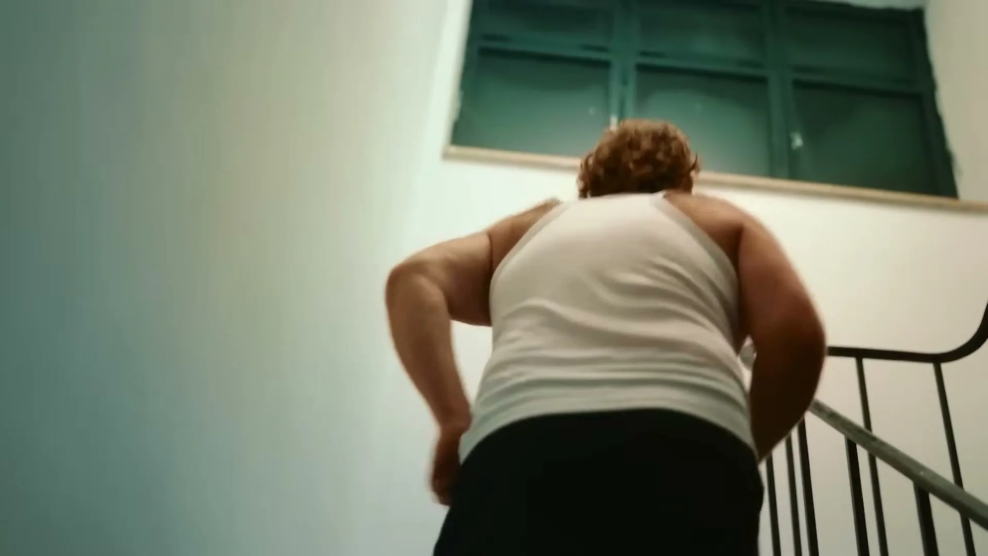 A man with curly hair and a white tank top walks up a flight of stairs in an apartment building.