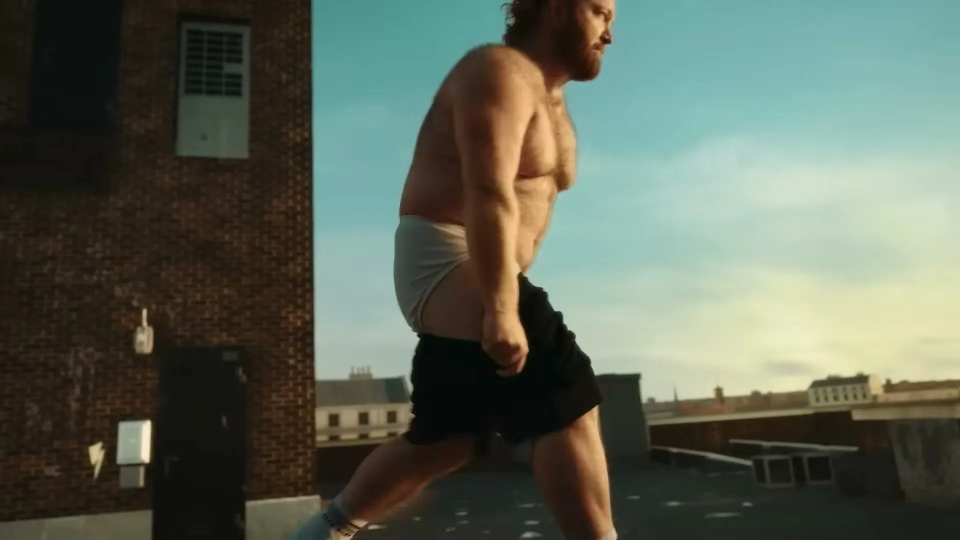 A shirtless, bearded man in his 30s, wearing black shorts and white underwear, walks on a rooftop under a clear sky.