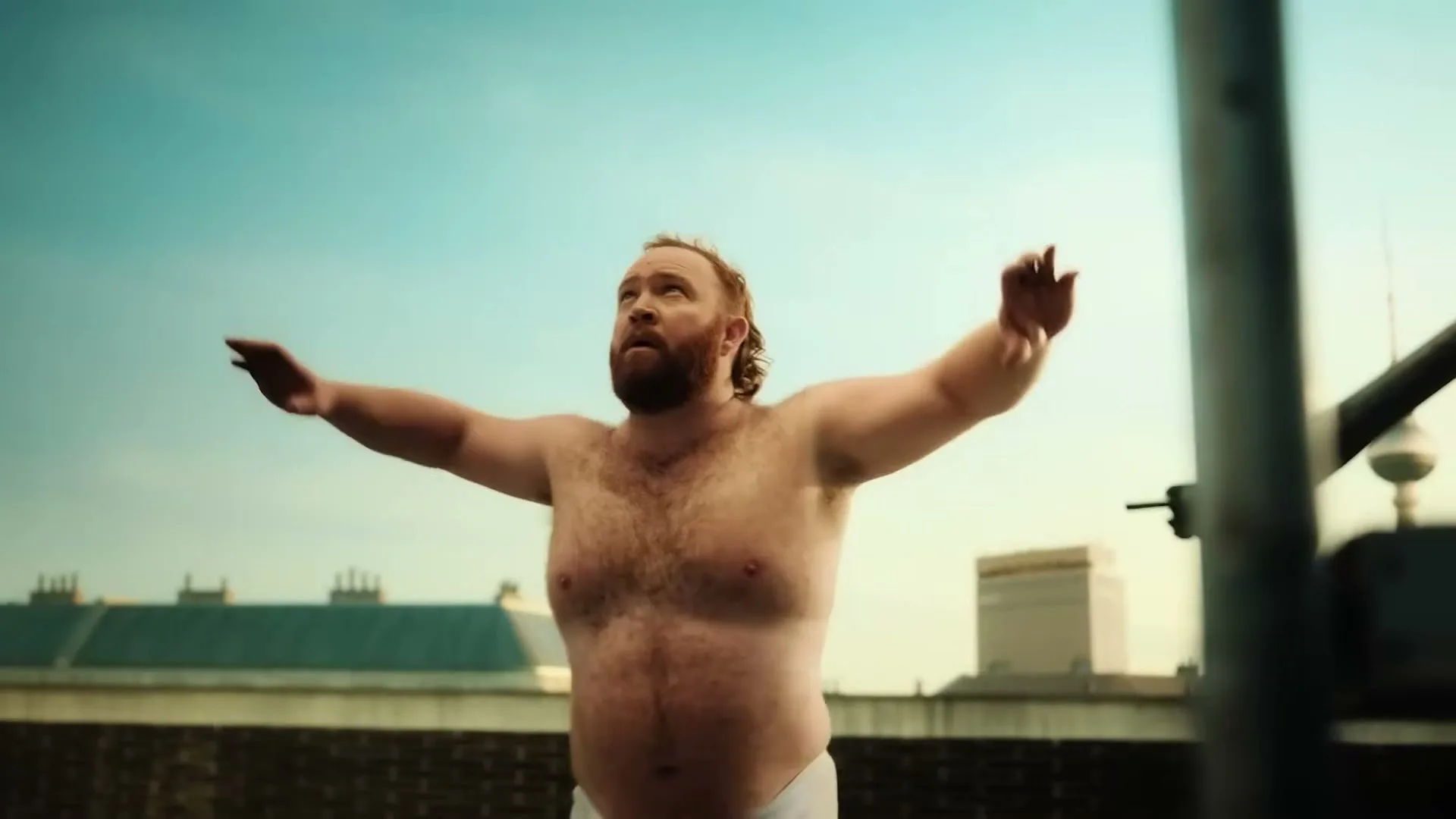 A shirtless, bearded man in white underwear stretches his arms upwards on a rooftop under a clear blue sky.