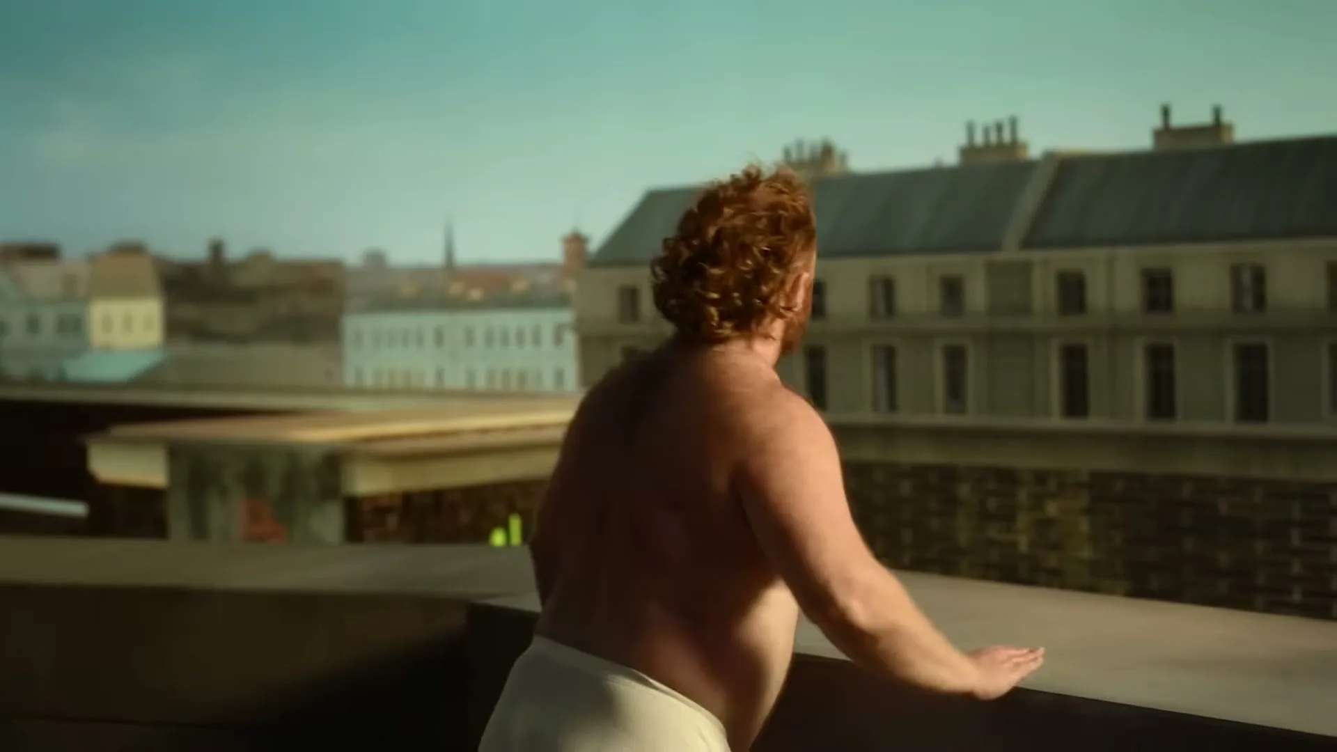 A shirtless, red-haired man in white underwear looks out over a city from a rooftop.