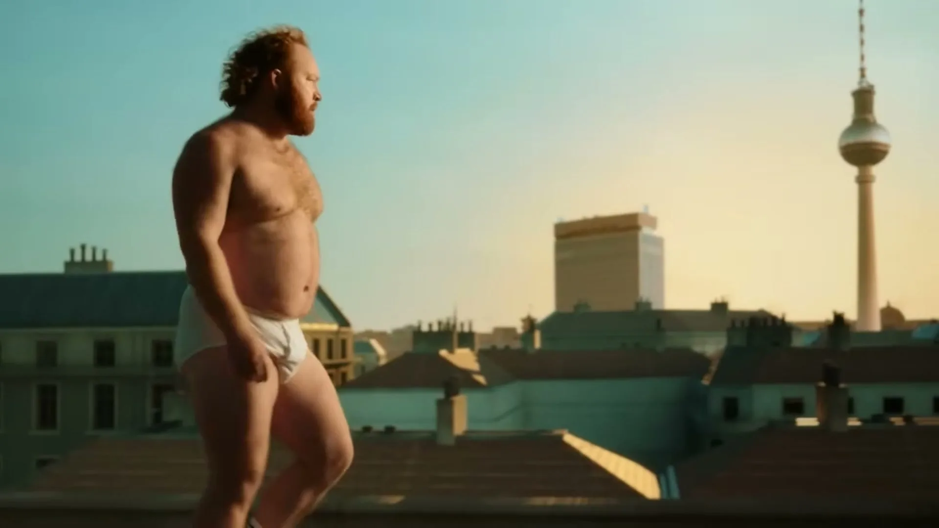 A shirtless, bearded man in white briefs walks across a rooftop with the Berlin TV Tower in the background.