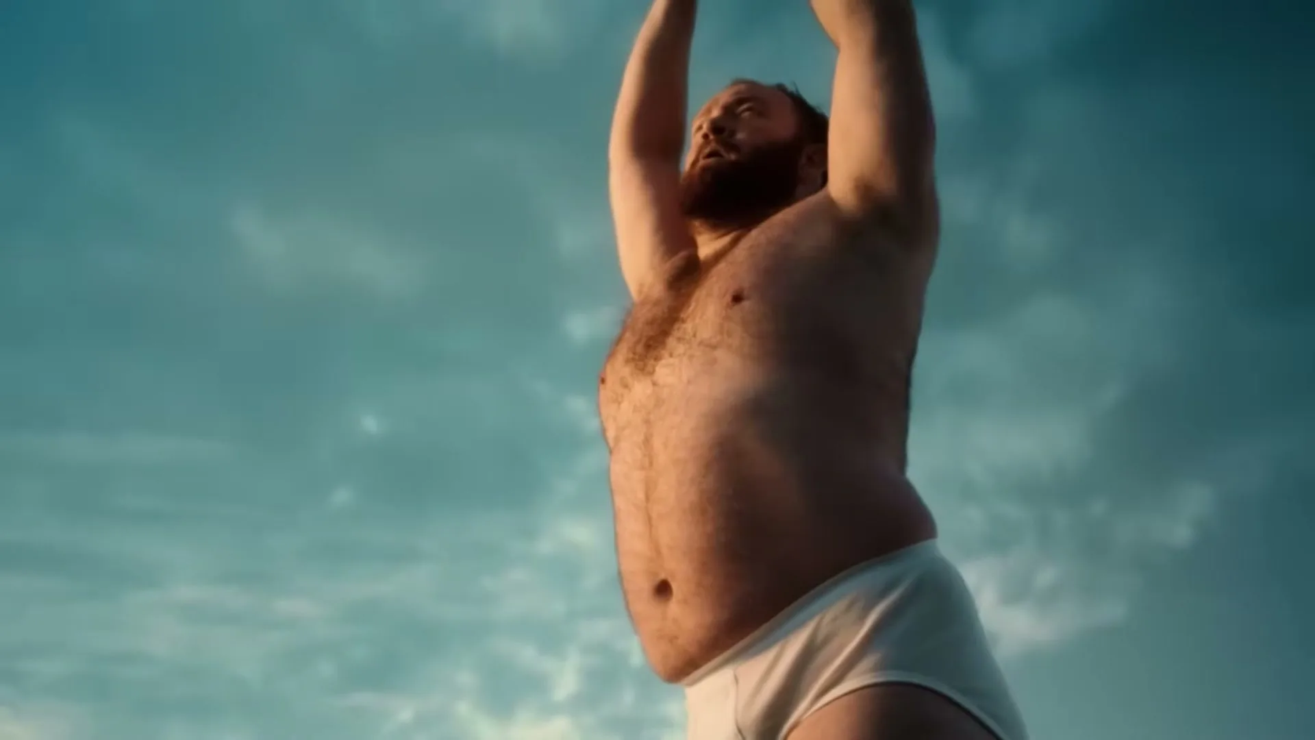 A hairy, overweight man in white briefs stretches his arms above his head against a blue sky.