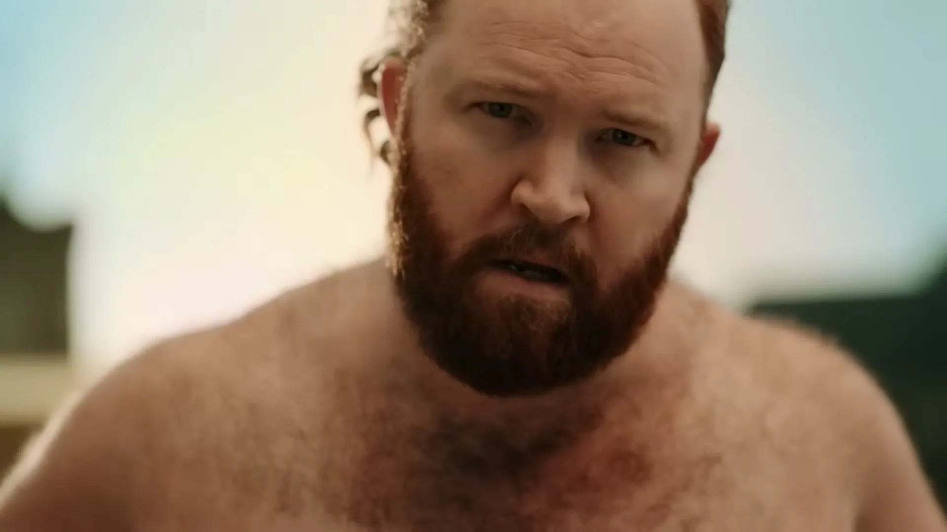 A shirtless, red-haired man with a beard looks directly at the camera with a confused expression in an outdoor setting.