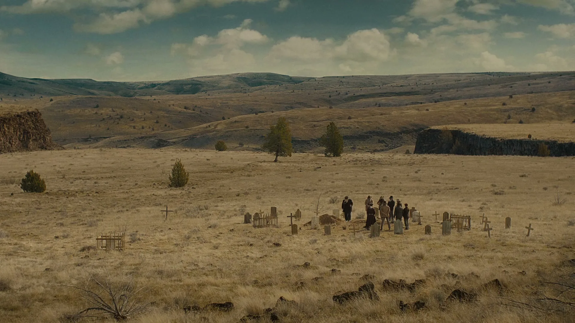 A group of people in old-fashioned clothing gather around a grave in a desolate, dry cemetery in a wide shot of a western landscape.