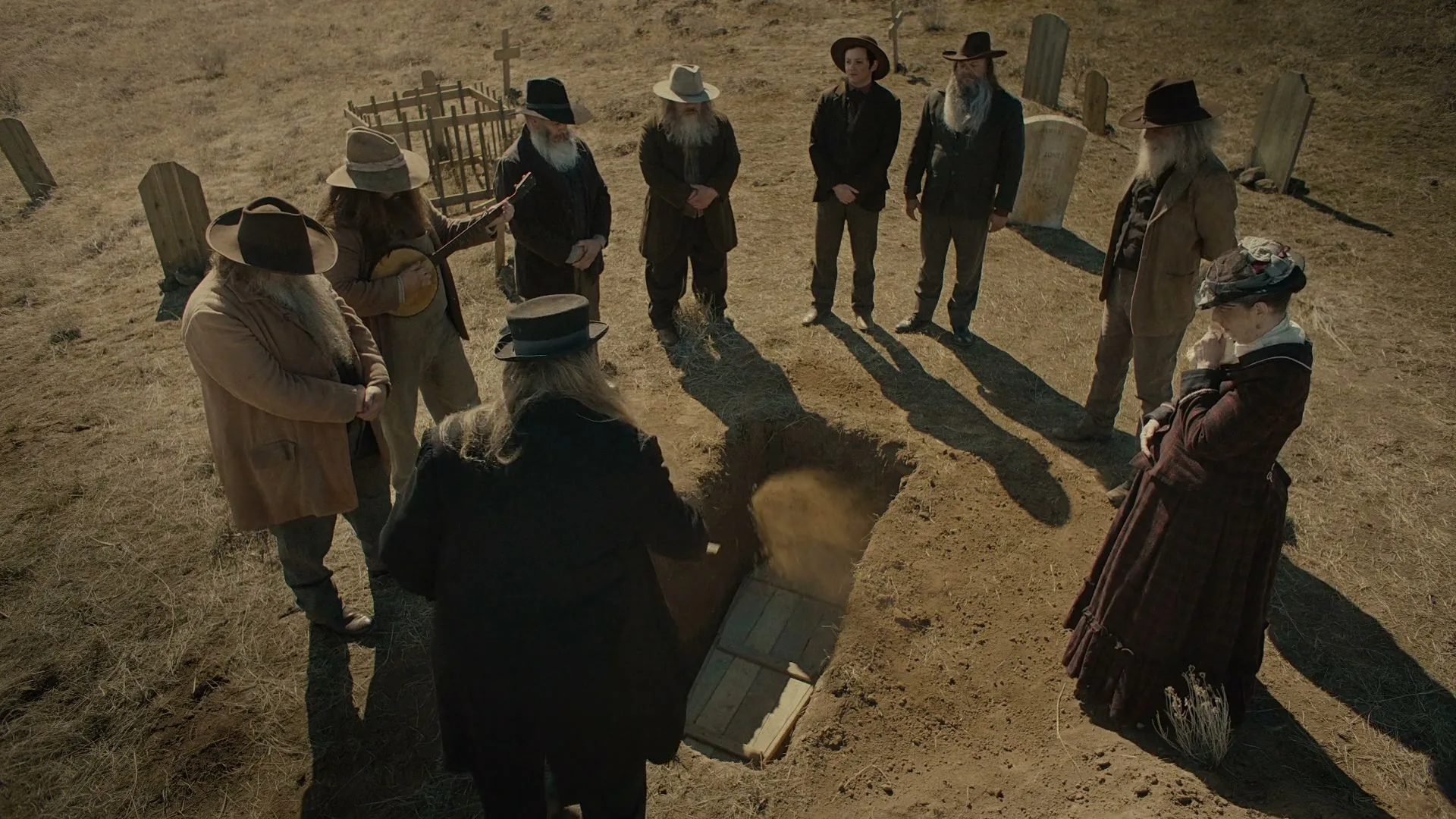 A group of men with long beards and a woman in period clothing stand around an open grave in a dusty cemetery, as a coffin is lowered.