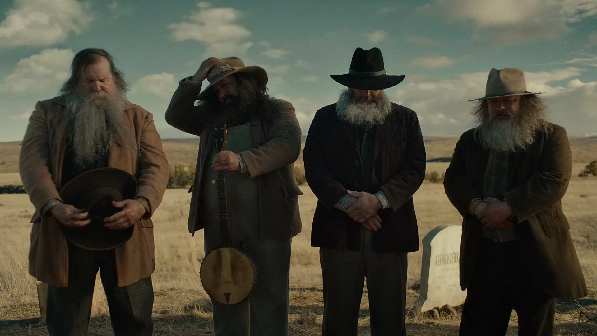Four men with long beards and hats stand in a row in a dry, grassy field with a tombstone visible.