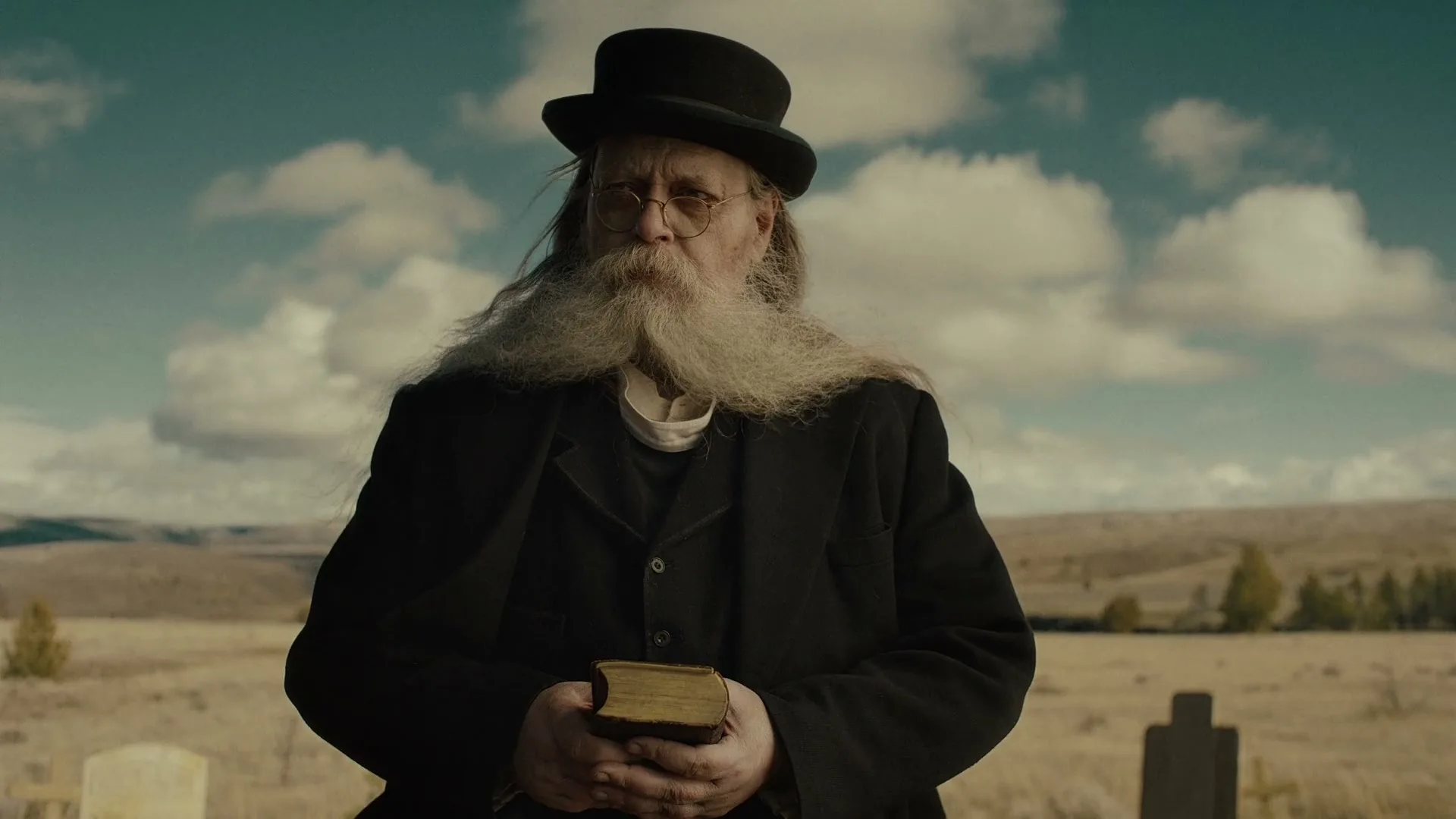 A man with a long white beard and a black bowler hat holds a book and speaks solemnly in a cemetery under a cloudy sky.