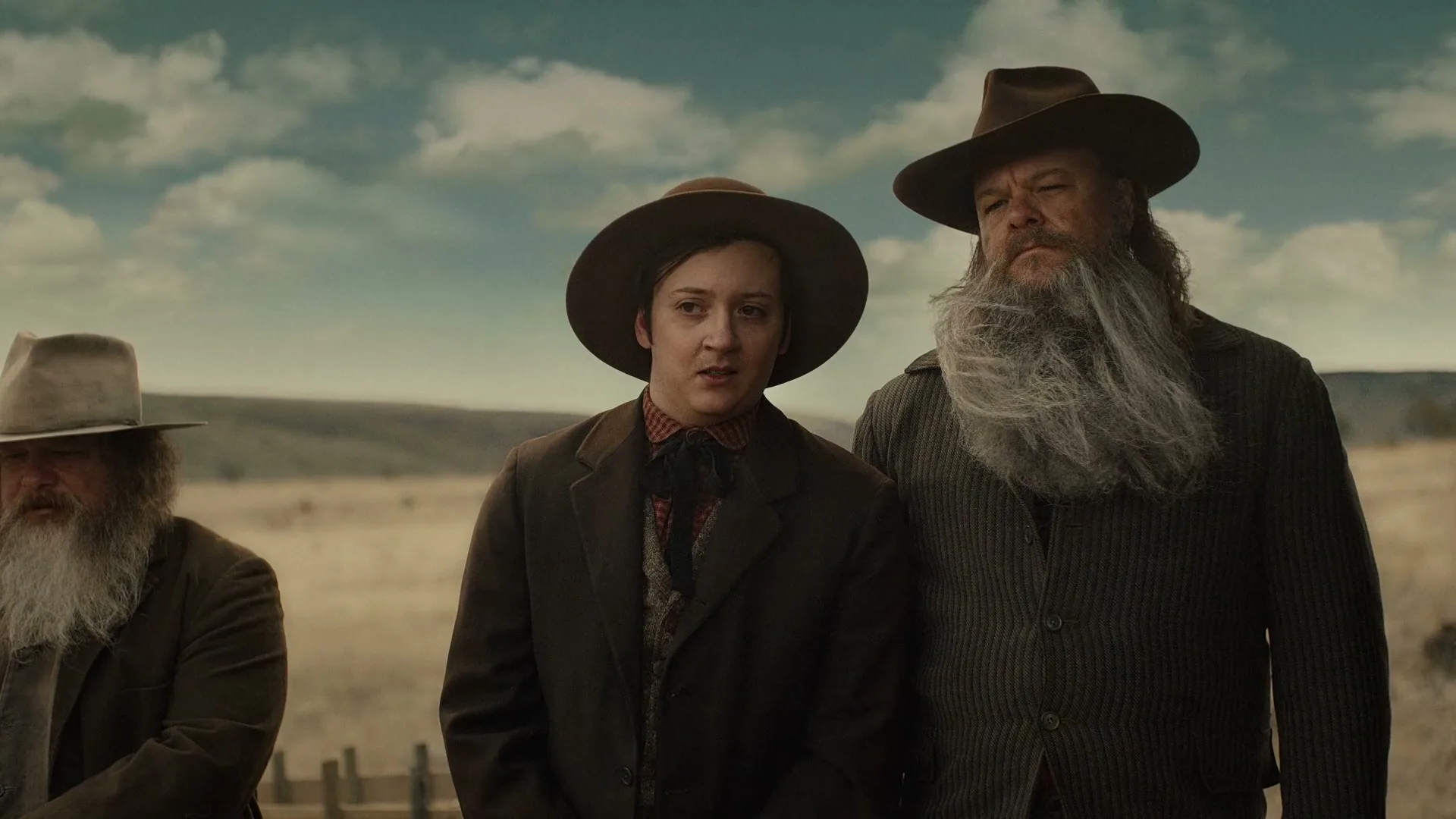 Three men with long beards and hats stand in a field under a cloudy sky, with the wind blowing their beards.