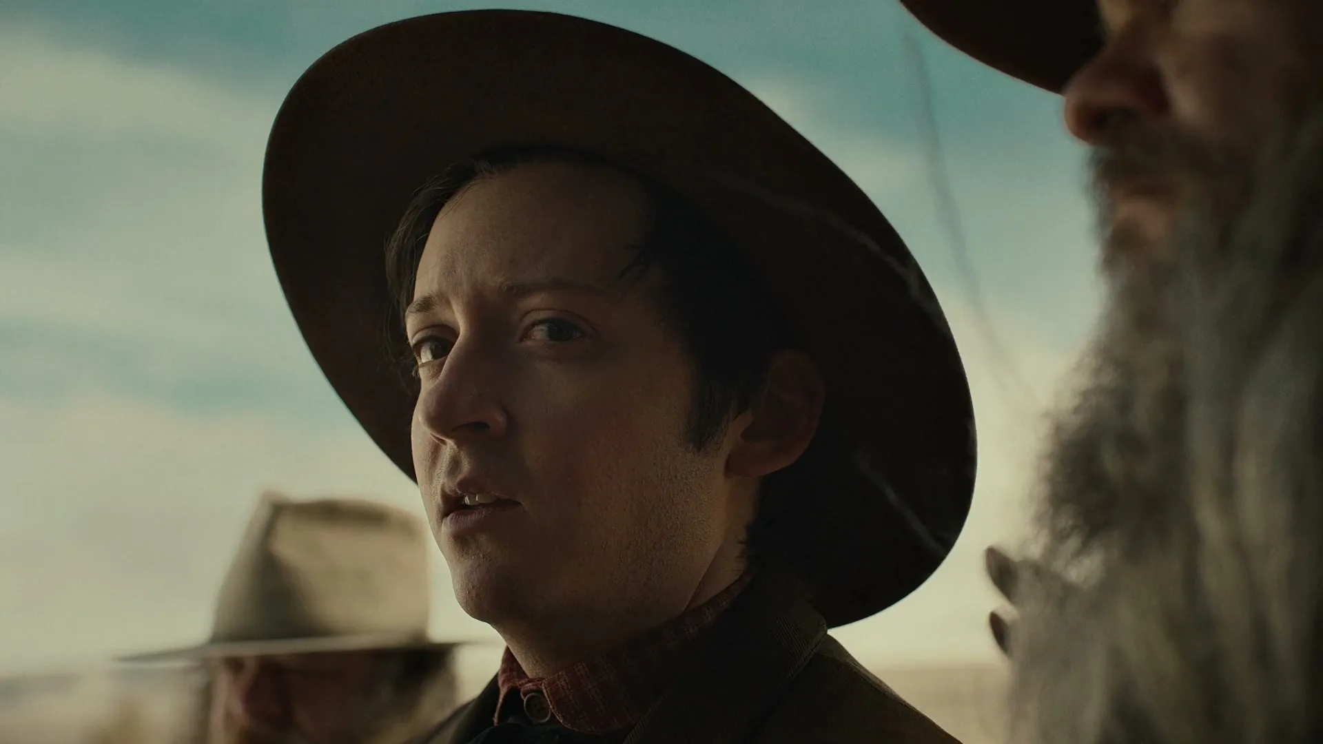 A young man in a cowboy hat looks concerned as an older man's hand rests on his shoulder in a dusty, outdoor setting.