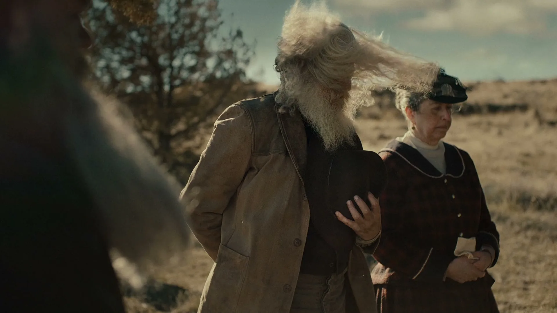 A man with a long white beard and hair, holding a hat, stands next to a woman in a dress, both looking concerned in a dry, outdoor setting.