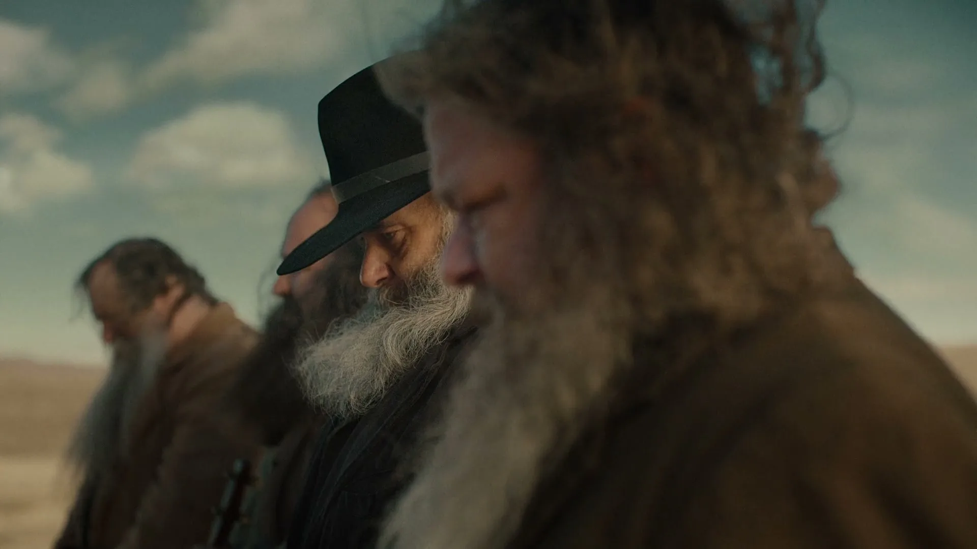 A group of men with long beards and hats stand in a line, looking down, in a desert landscape under a cloudy sky.