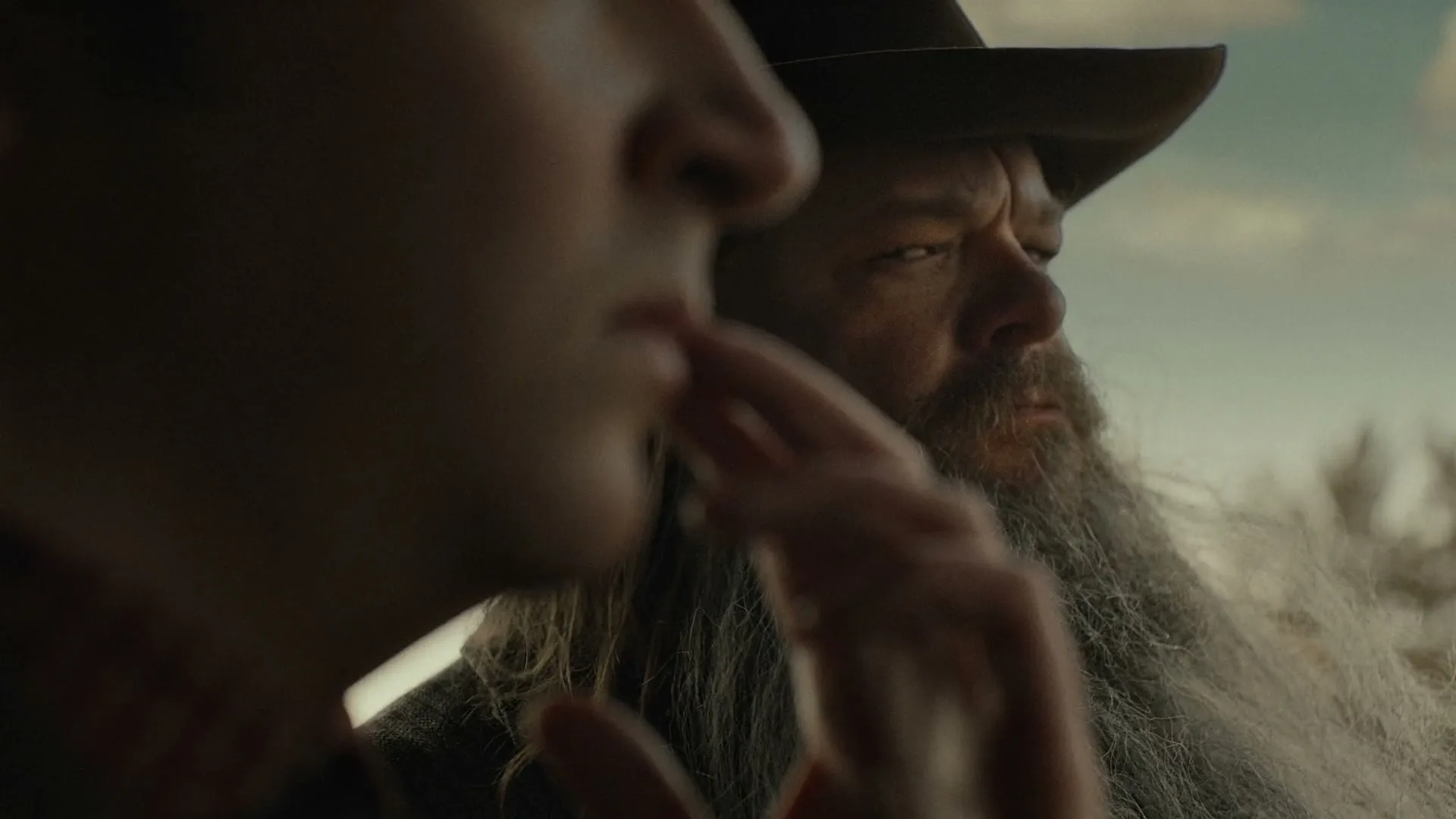 A young man with a finger to his lips looks forward, while an older man with a long beard and cowboy hat looks out a window in a vehicle.