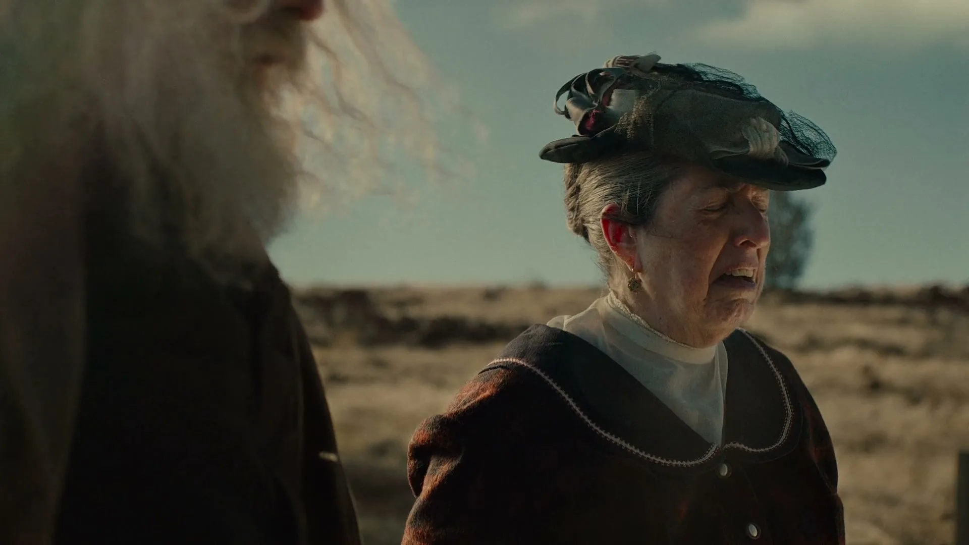 An older woman in a period dress and hat cries out in distress next to an older man with a long white beard in a dry, outdoor setting.