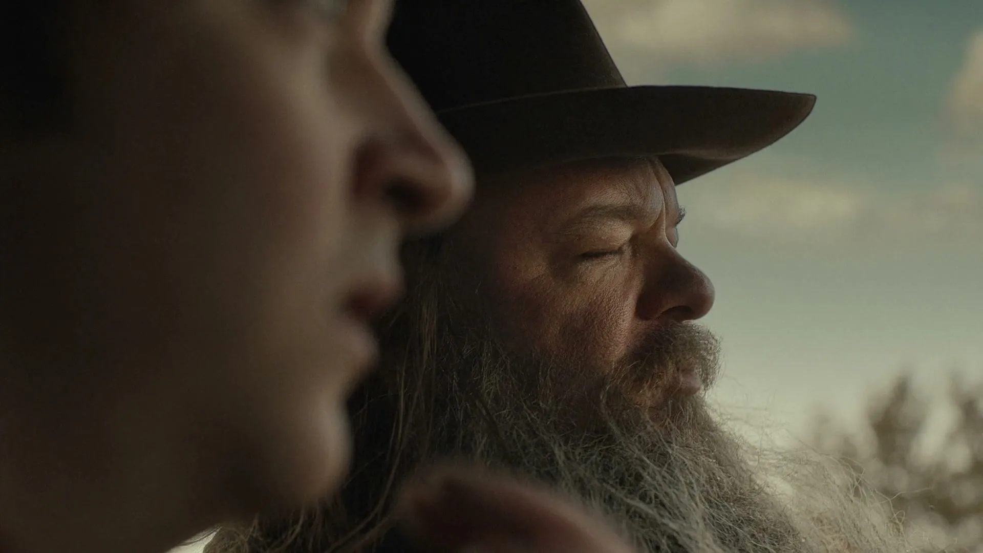 A young man and an older man with a long beard and cowboy hat look off into the distance, with a cloudy sky in the background.