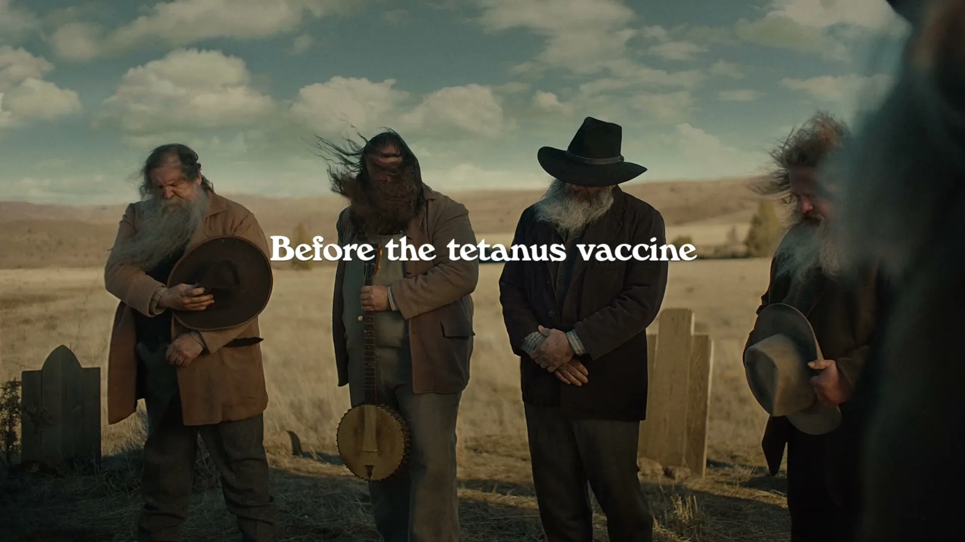 Four men with long beards and hats stand in a windy, desolate cemetery under a cloudy sky, with text appearing on screen.