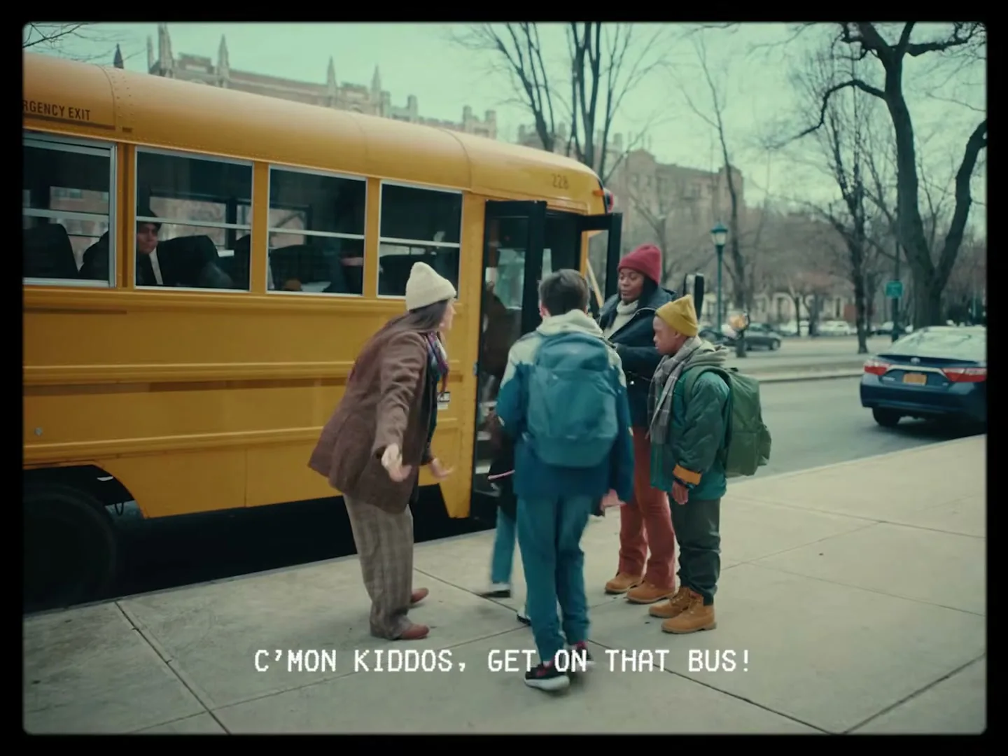 A woman in a brown coat and white beanie gestures towards a yellow school bus as children with backpacks board it on a city street.