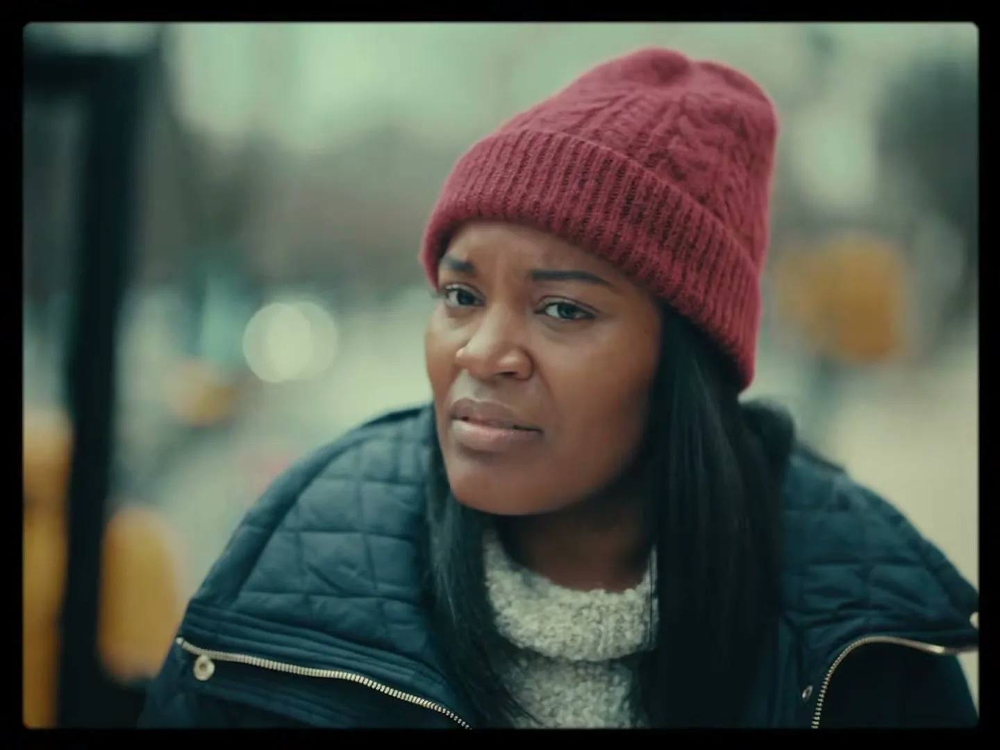 A young Black woman in a red beanie and quilted jacket looks confused and slightly disgusted, outdoors on a city street.