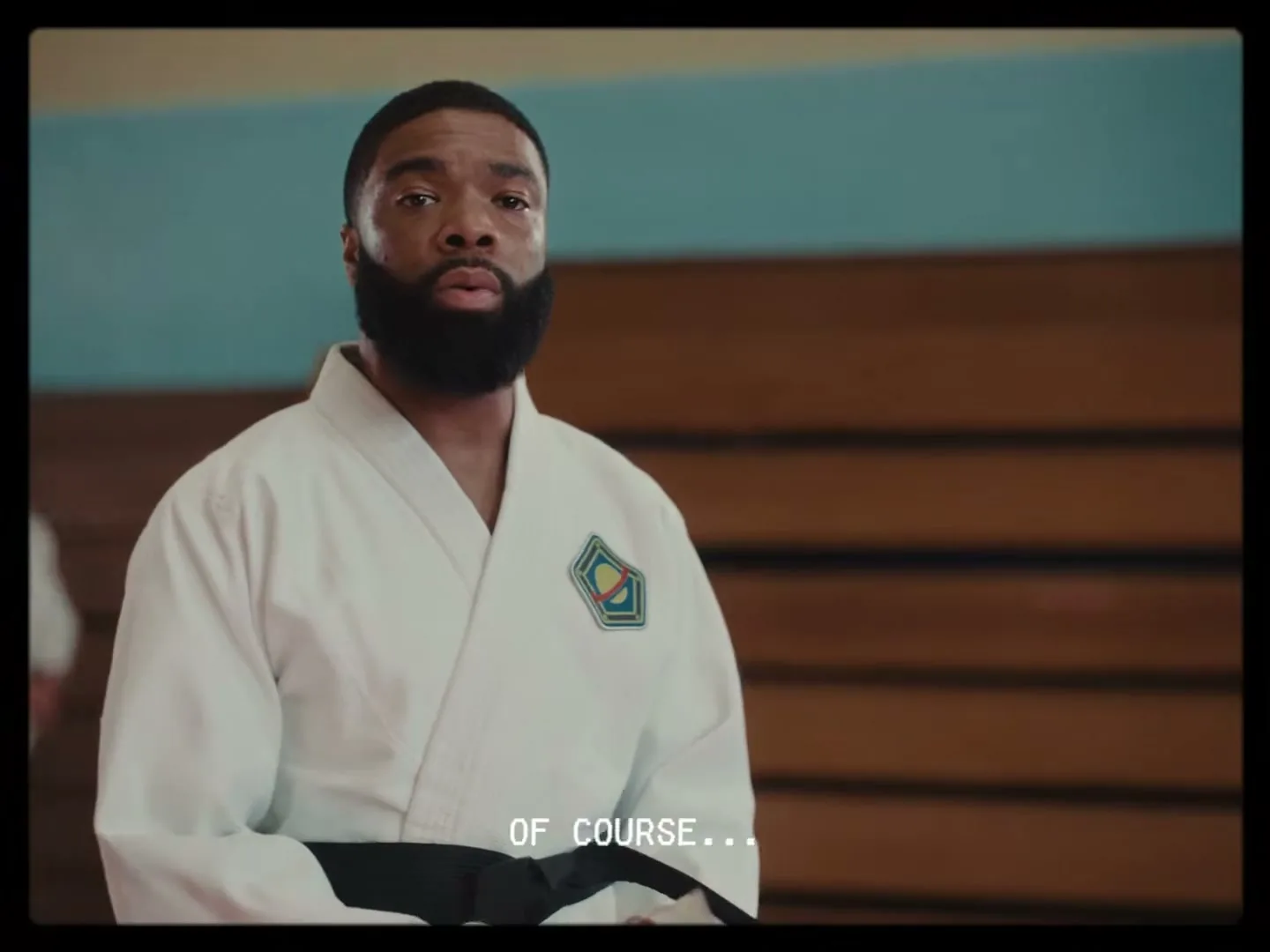 A Black man with a beard in a white karate gi with a black belt looks at the camera, then down at a book in a dojo.
