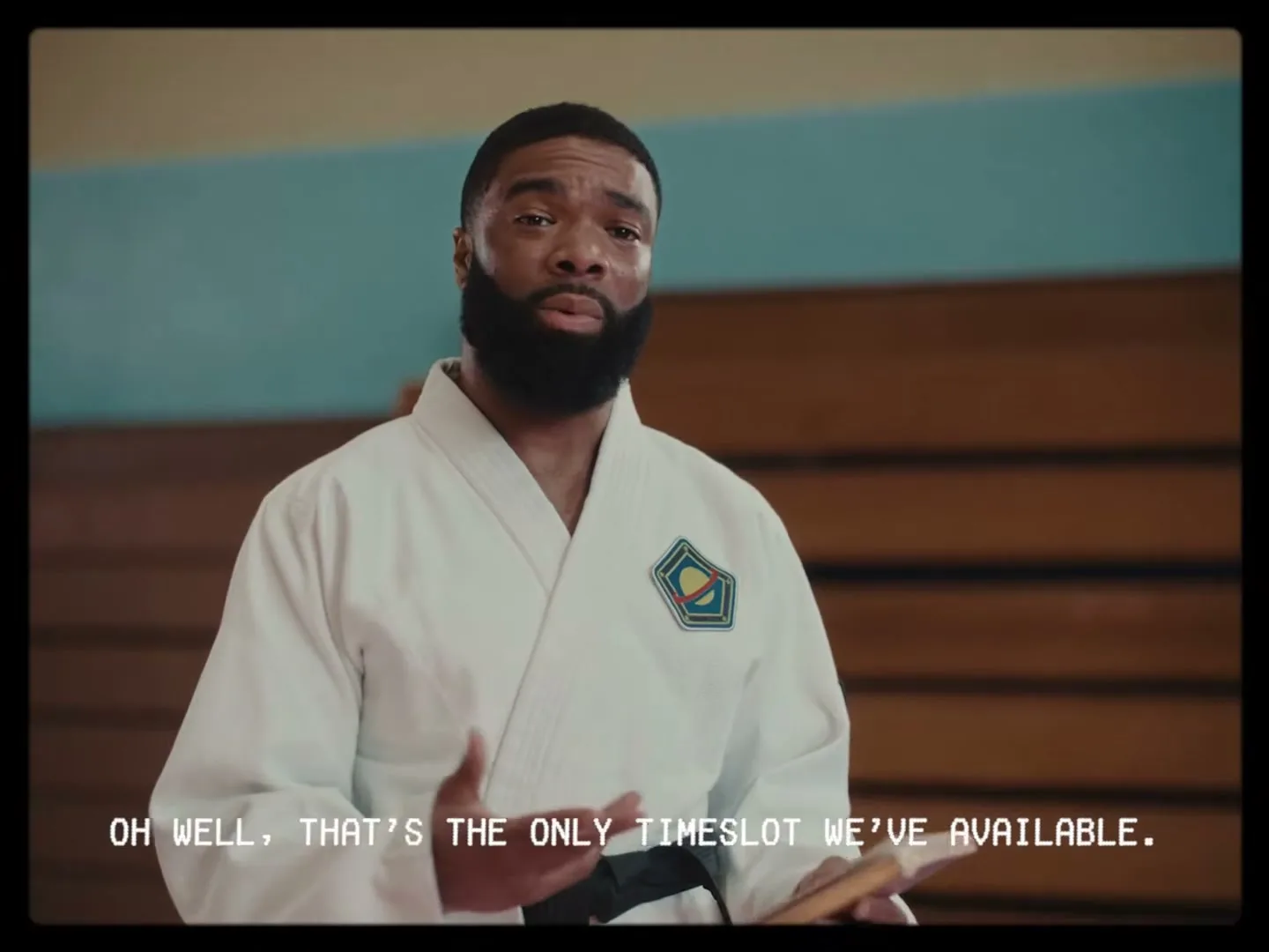 A Black man with a beard, wearing a white karate gi, stands in a gym and speaks while holding a book.
