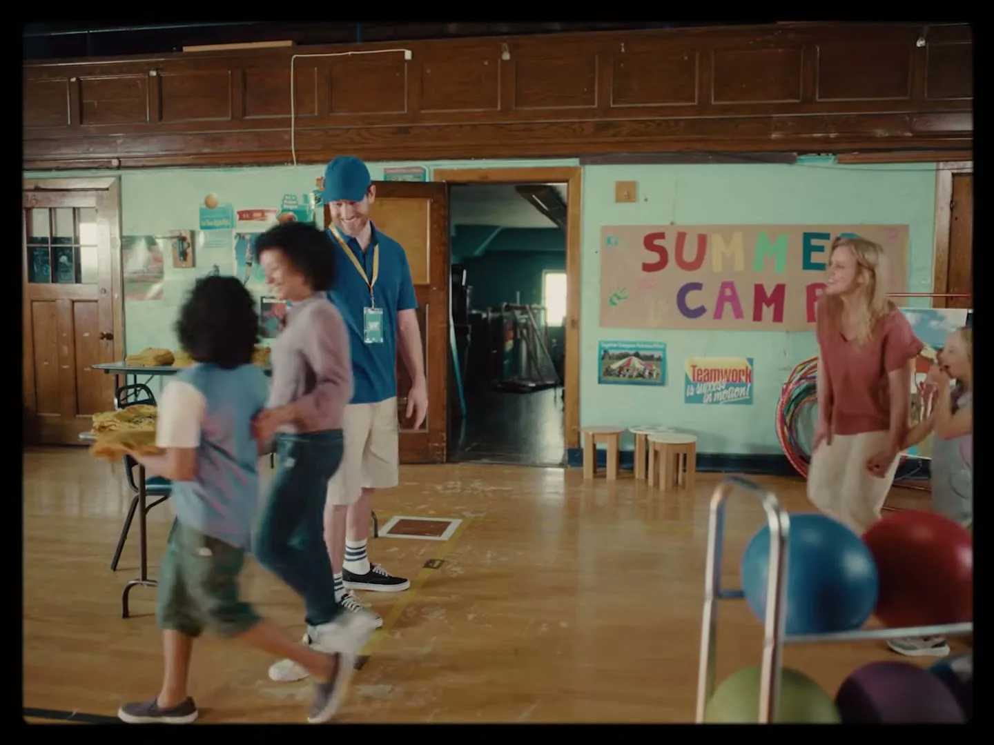 A male camp counselor in a blue shirt and hat smiles as children and a female counselor walk past him in a school gym during summer camp.