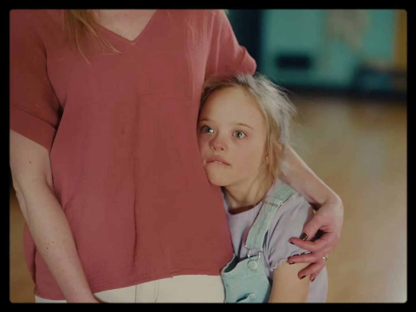 A young girl with Down syndrome rests her head on an adult woman's torso, looking up with a worried expression in an interior setting.