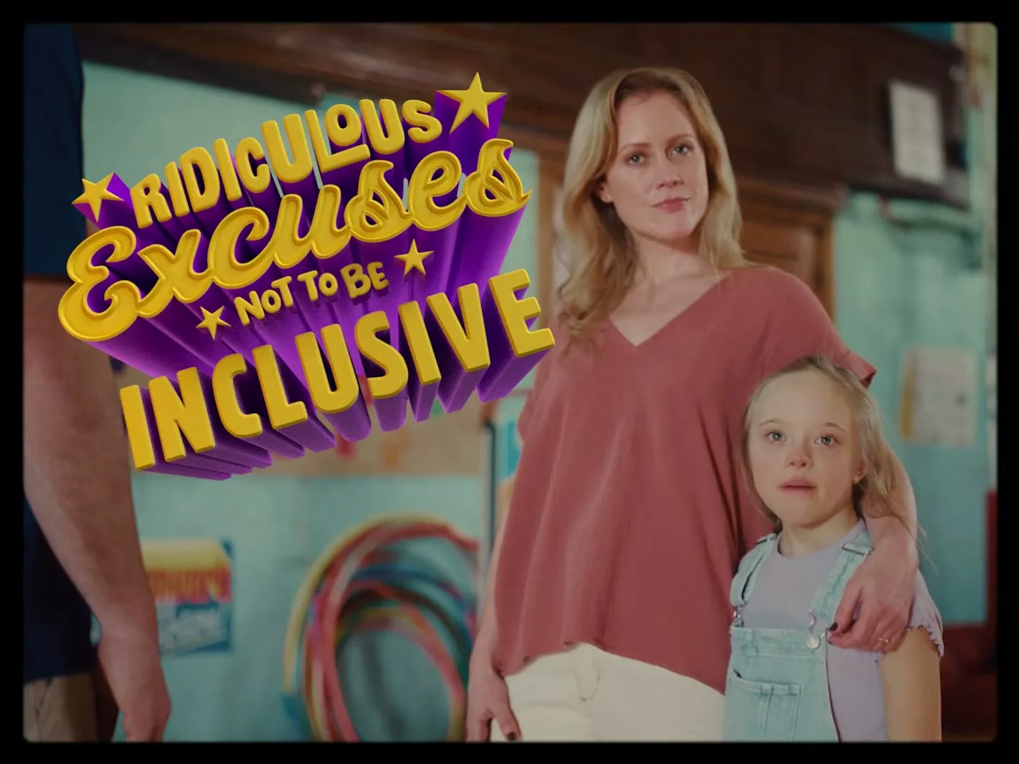 A woman with blonde hair and a young girl with Down syndrome stand together, looking at the camera with a serious expression in a brightly lit room with a "Summer Camp" sign.
