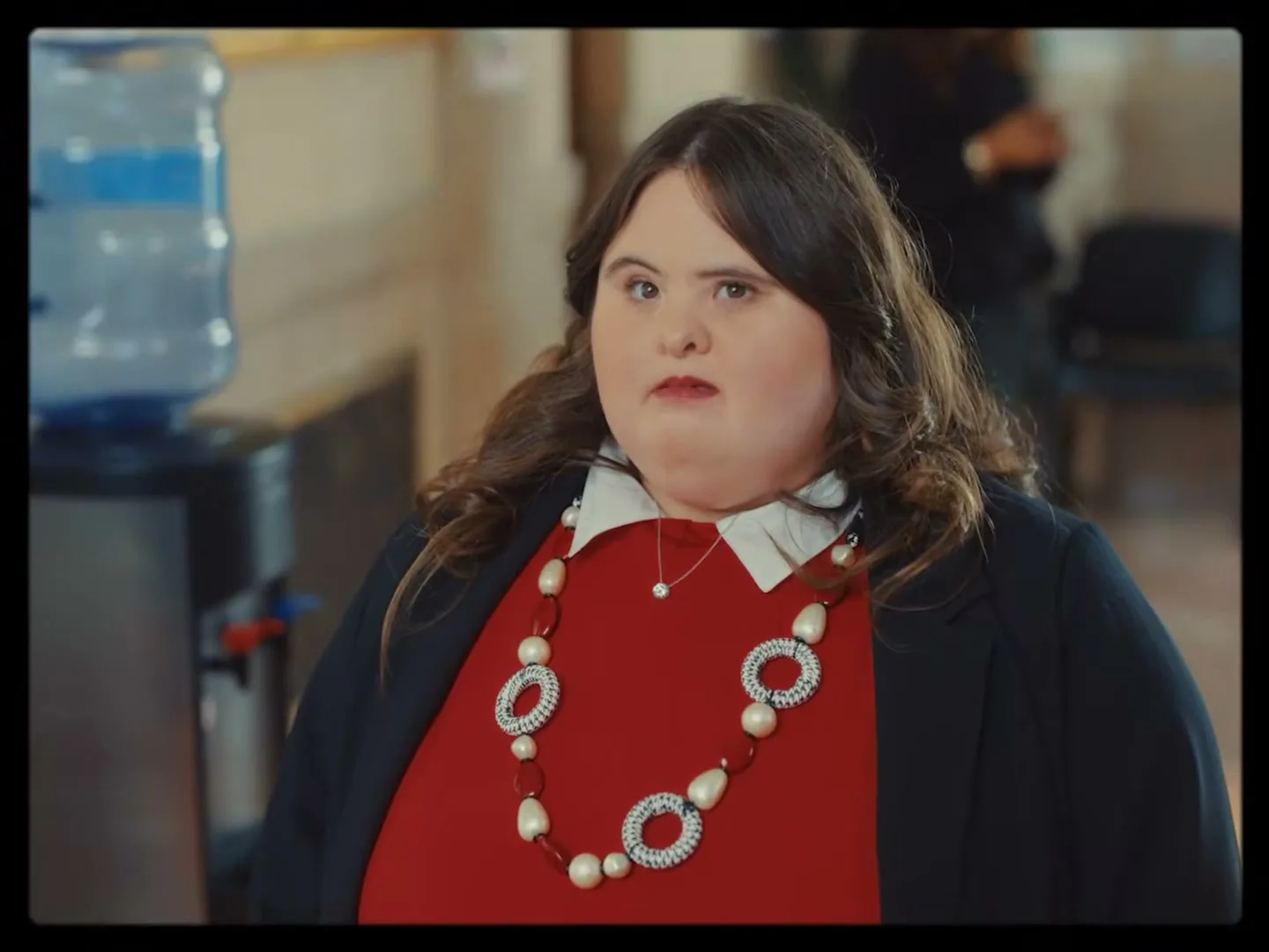 A woman with Down syndrome, wearing a red top and black blazer, stands in an office next to a water cooler, looking directly at the camera with a serious expression.