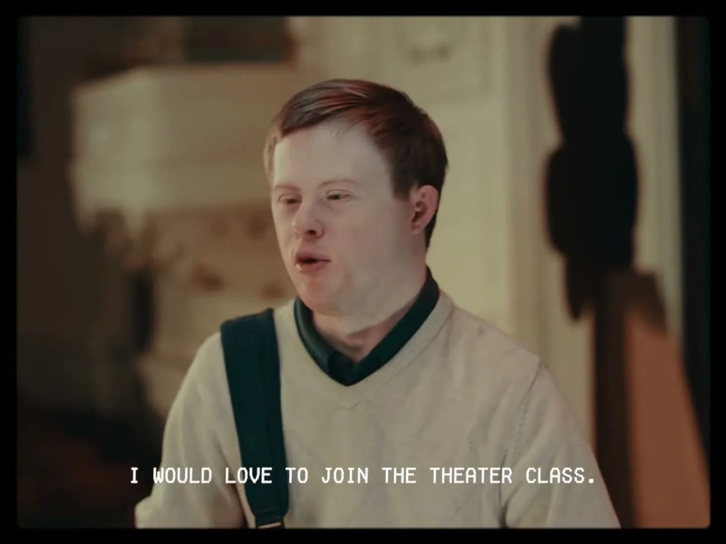 A young man with Down syndrome, wearing a cream sweater and green polo, speaks directly to the camera in an interior setting.