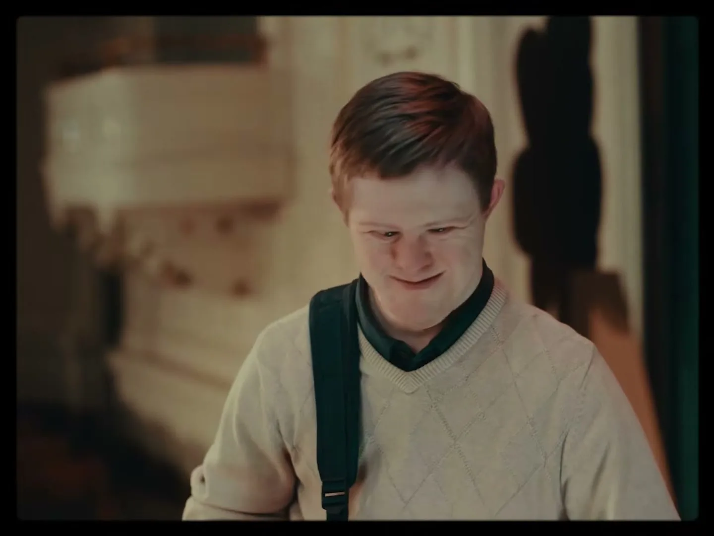 A young man with Down syndrome, wearing a cream sweater and a black strap over his shoulder, looks down and then turns to walk away in a building.
