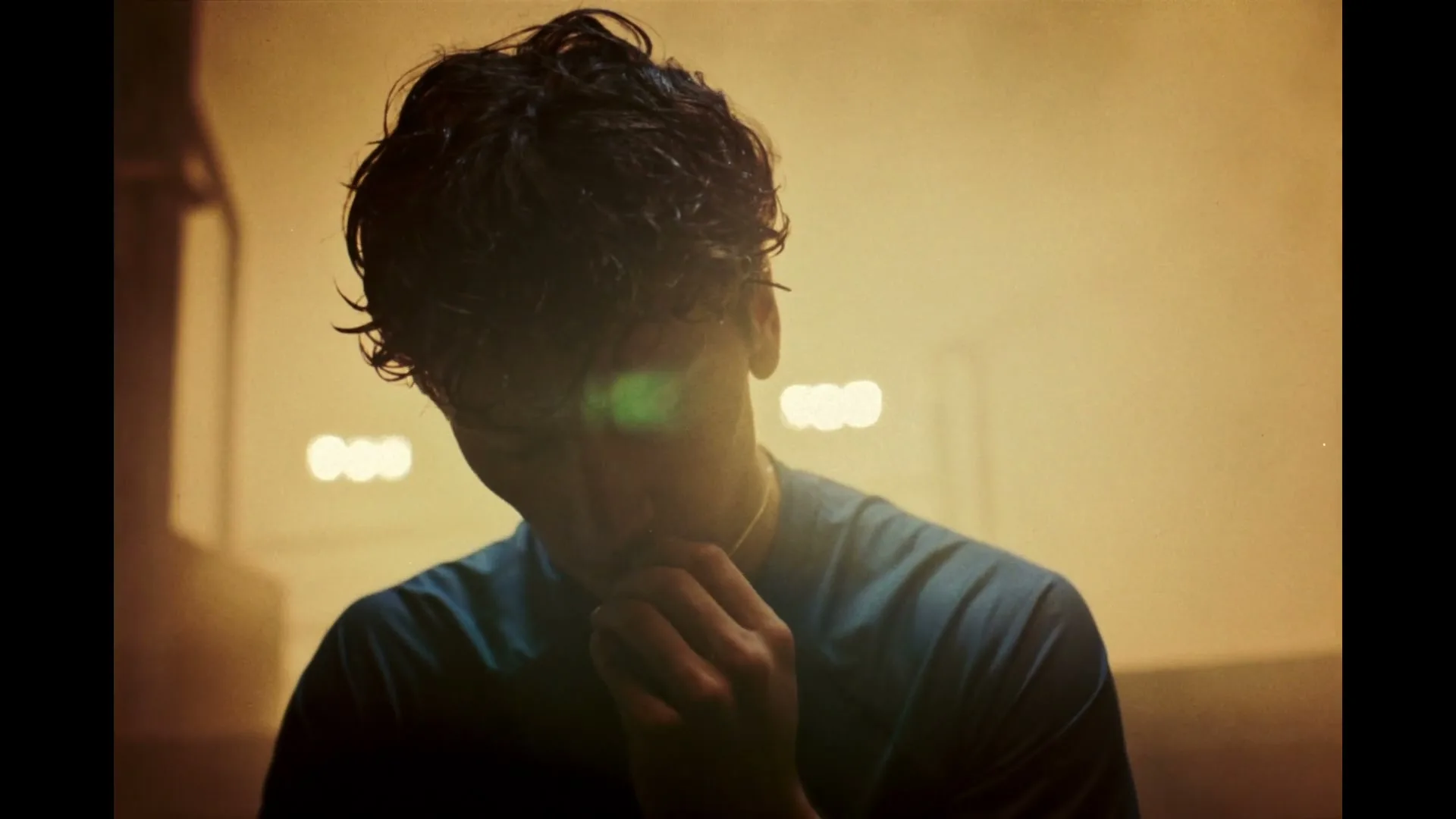 A young man with dark, curly hair, wearing a blue shirt, looks down with his hand near his chin in a dimly lit, hazy interior.