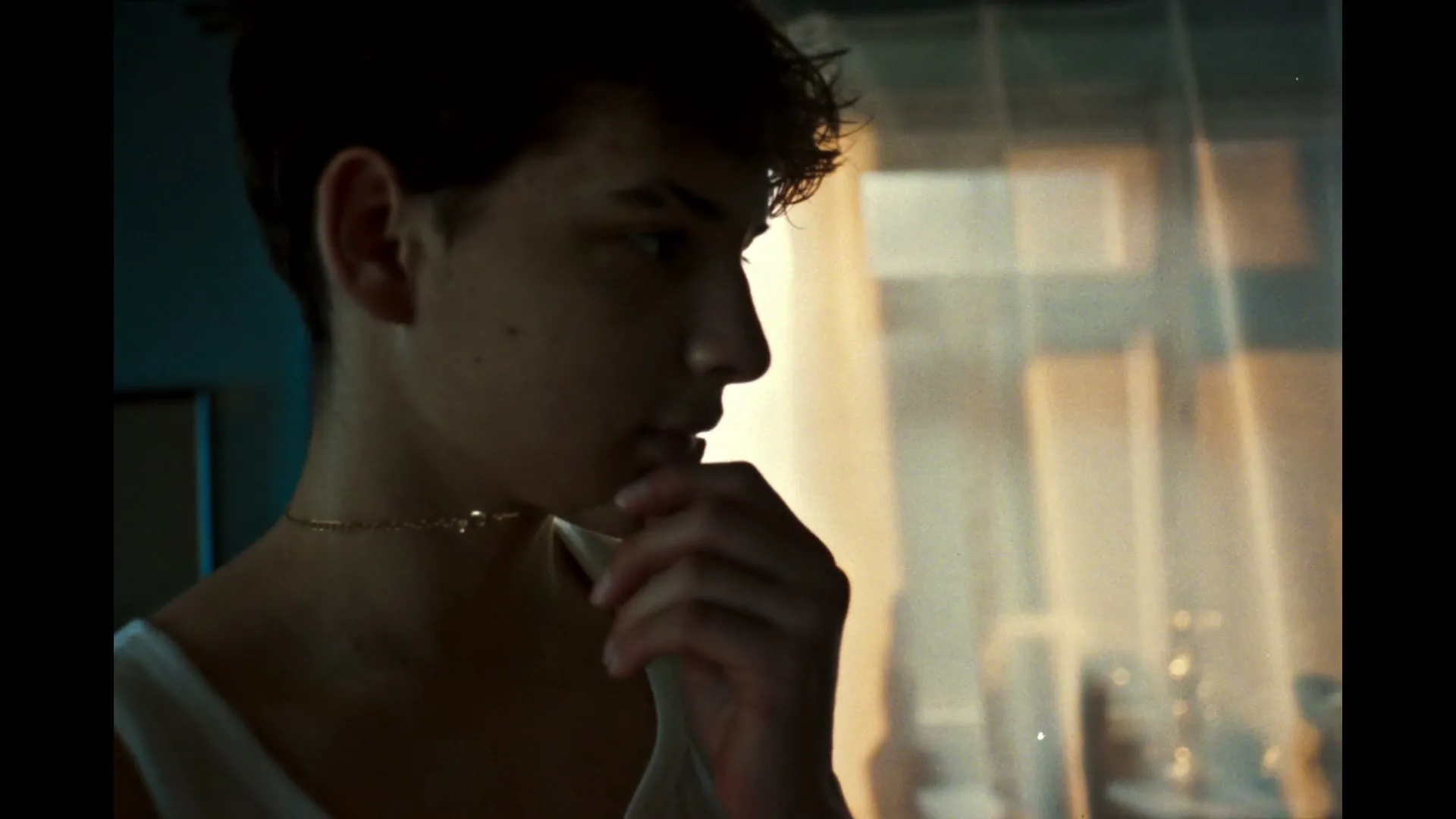 A young person with short dark hair and a gold chain looks contemplatively to the right, with soft light from a window in the background.