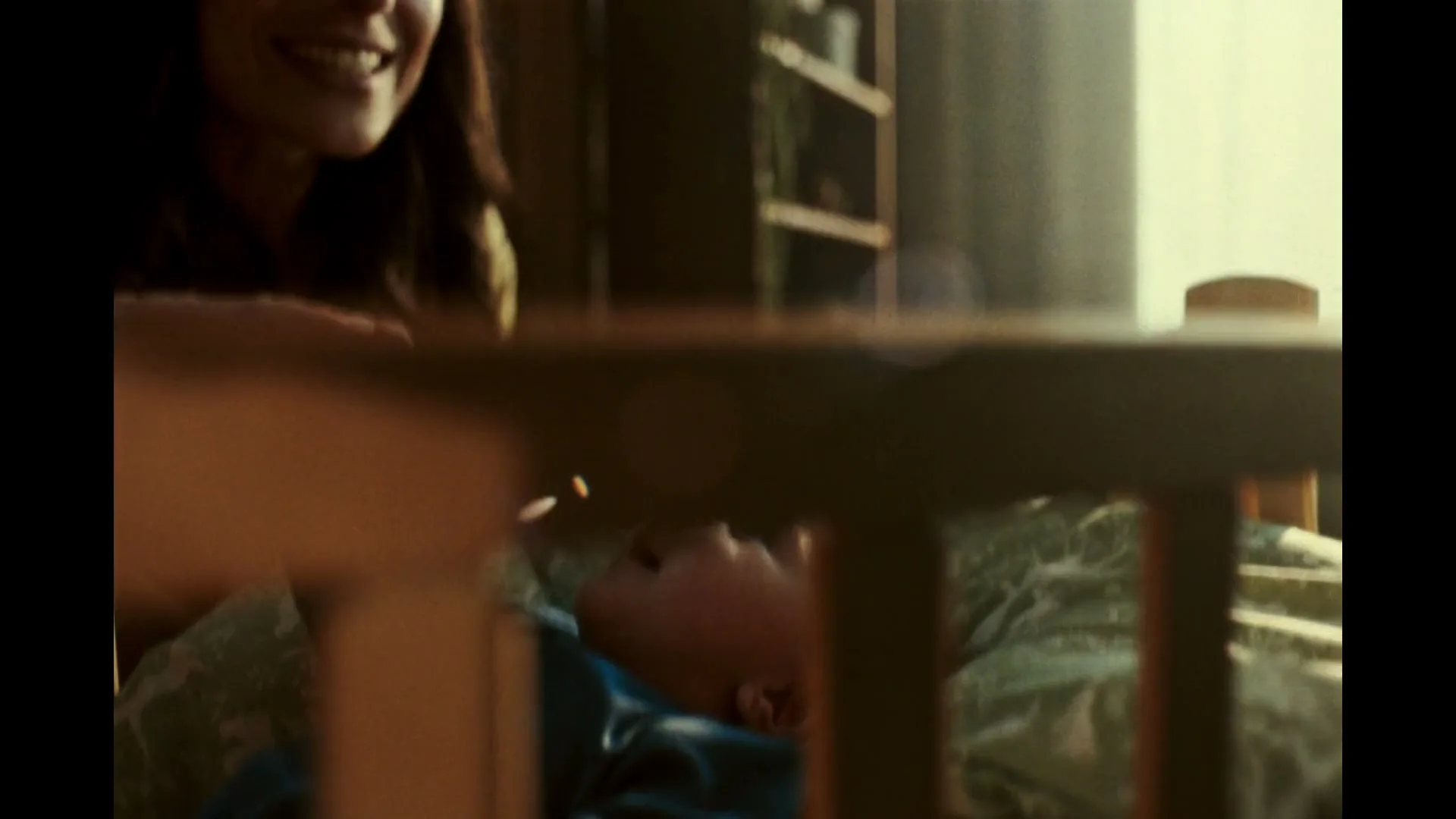 A baby in a crib looks up as a woman's hand gently touches its face, then the woman smiles at the baby in a cozy room.