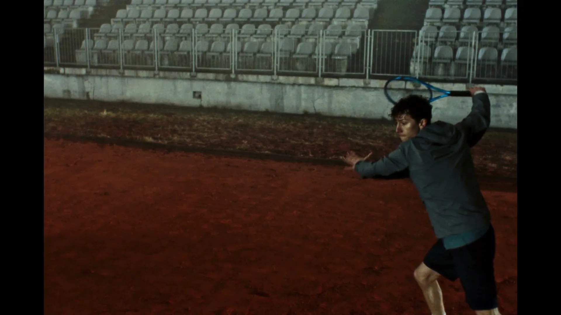 A male tennis player in a grey jacket and black shorts swings a tennis racket on a clay court at night, with empty stadium seats in the background.