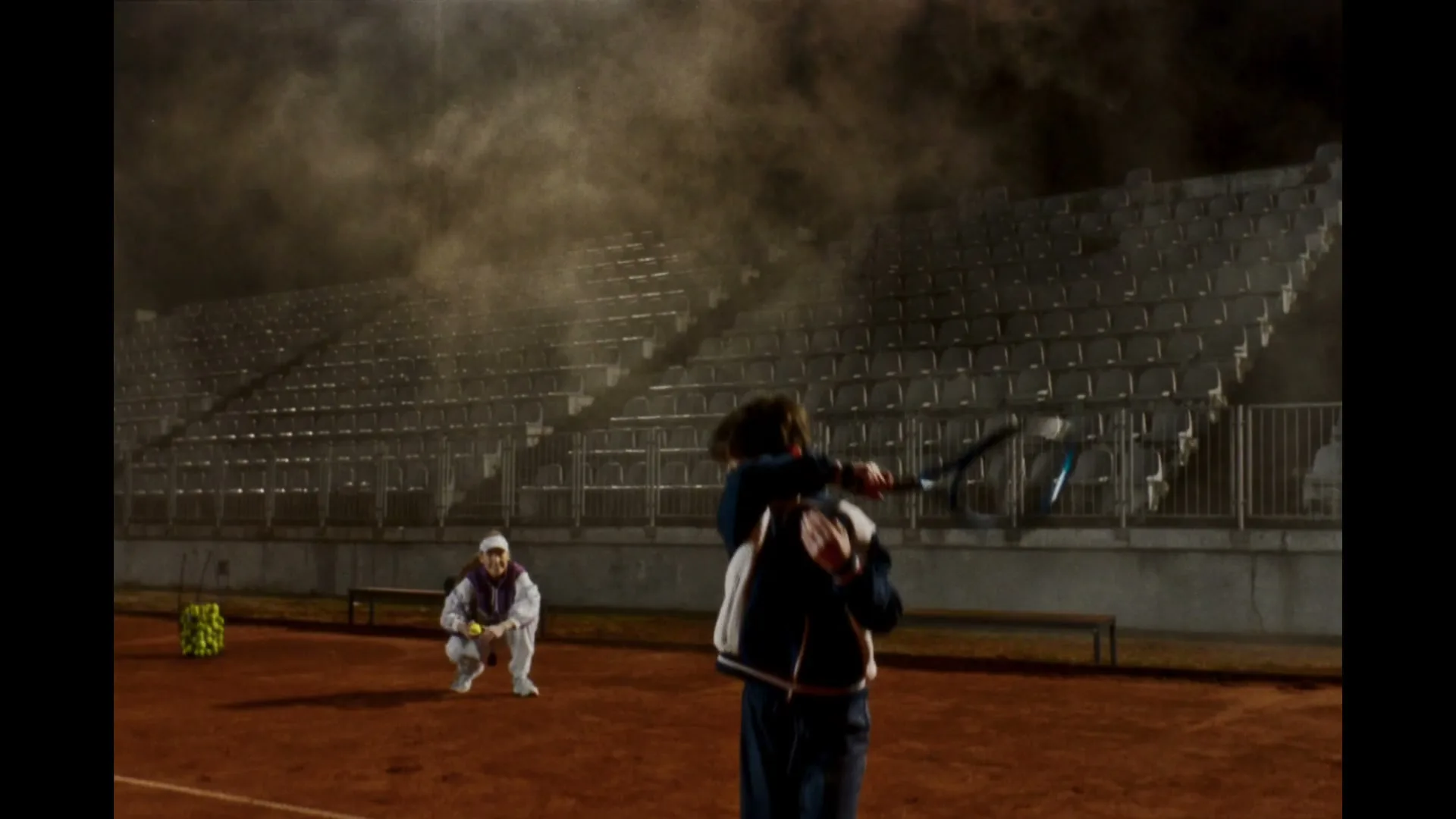A young boy in a tracksuit practices tennis on a clay court at night, while a coach crouches in the background.
